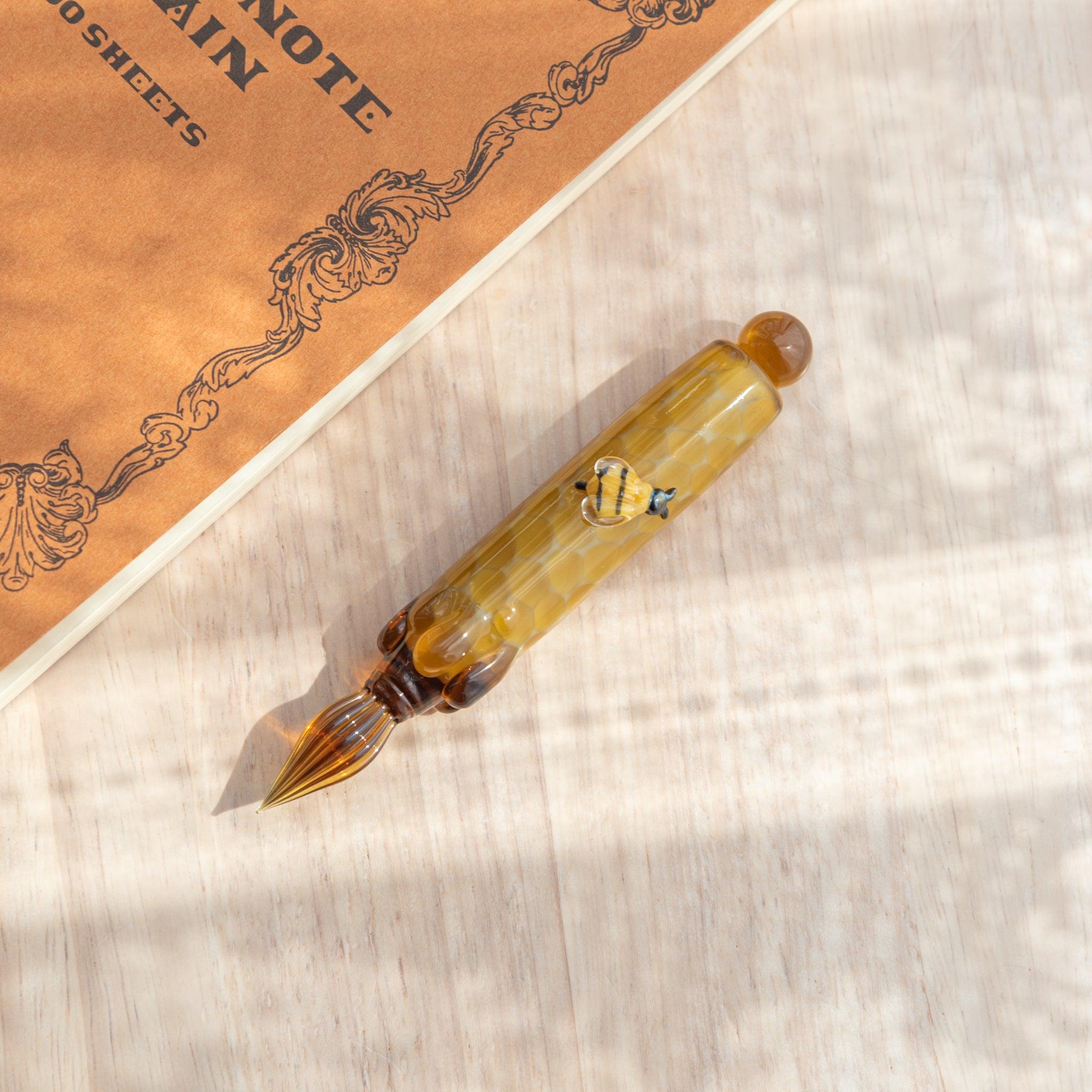 Top-down view of the Honey Bee Glass Dip Pen beside a notebook, with clear bee figure on yellow glass.