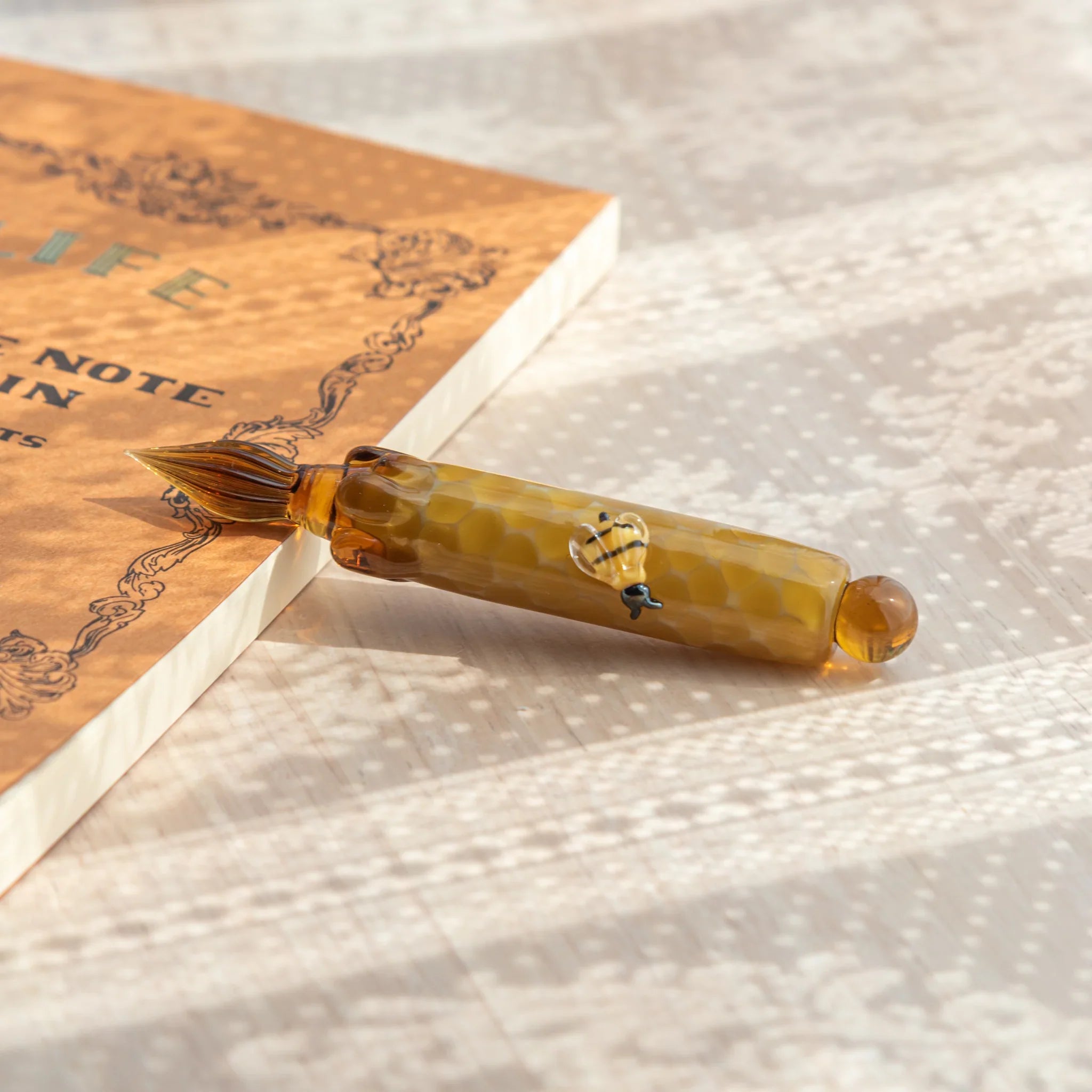 Japanese glass dip pen with amber honeycomb barrel, honey bee figure on body, multi-tine nib resting on a kraft notebook