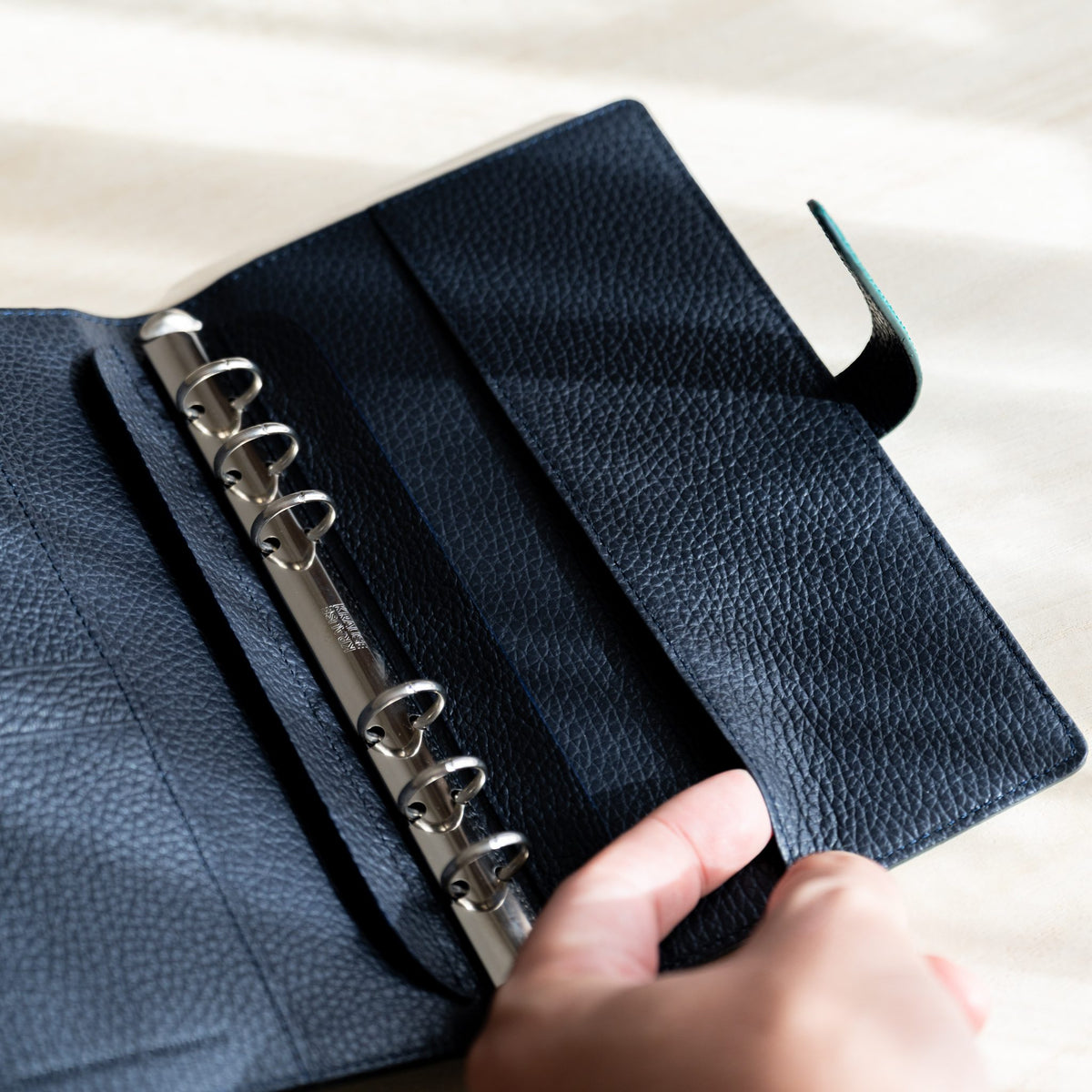 Fingers lifting the inner flap pocket of the navy leather lining next to the binder rings
