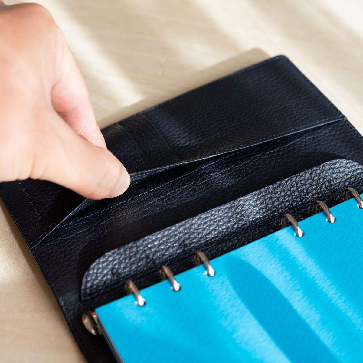 Fingers opening a pocket in the navy leather interior beside blue pages on the rings