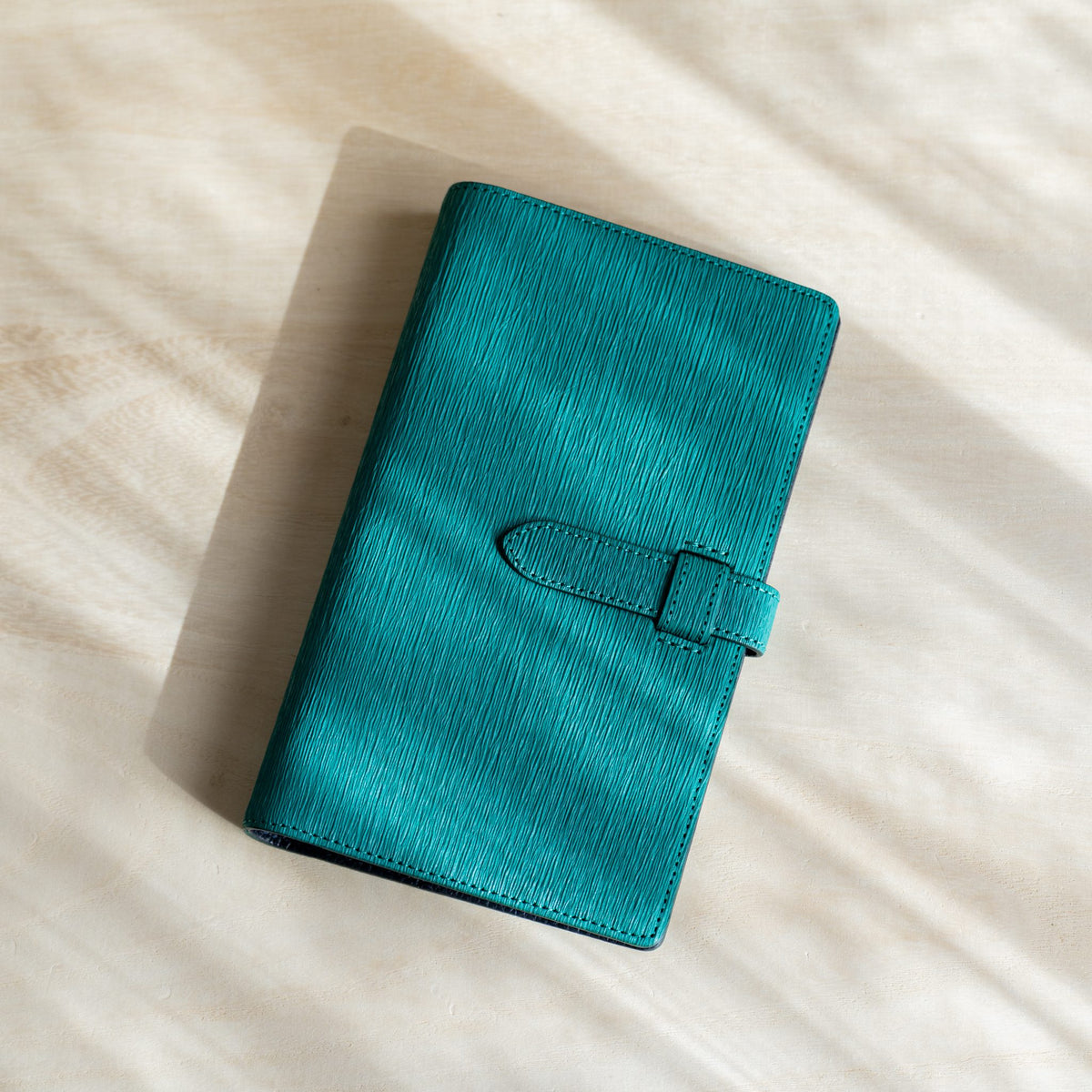 Teal Ise Gikakushi paper leather planner lying at an angle, vertical crease texture and strap closure