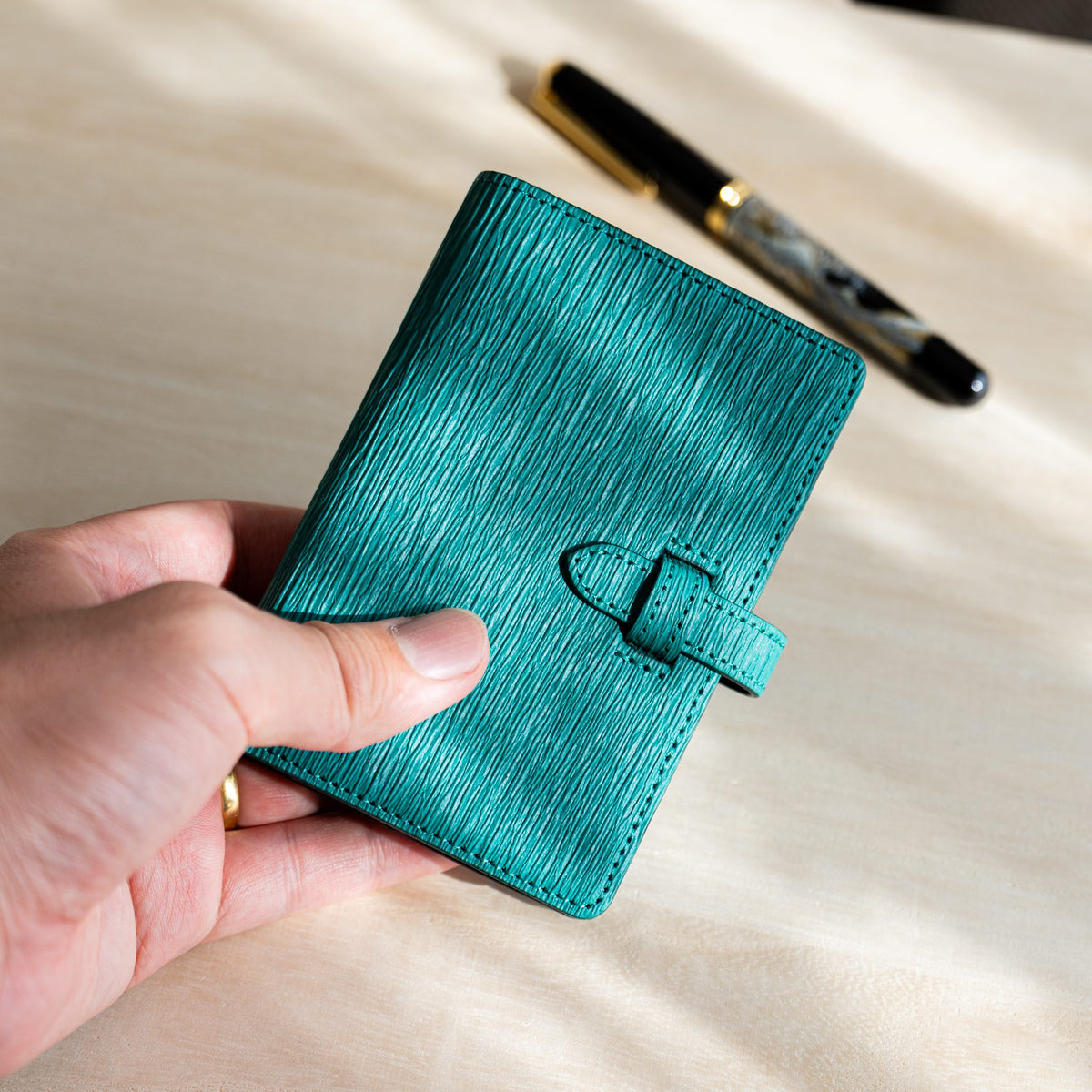 Hand holding the small teal Ise Gikakushi planner with a fountain pen in the background