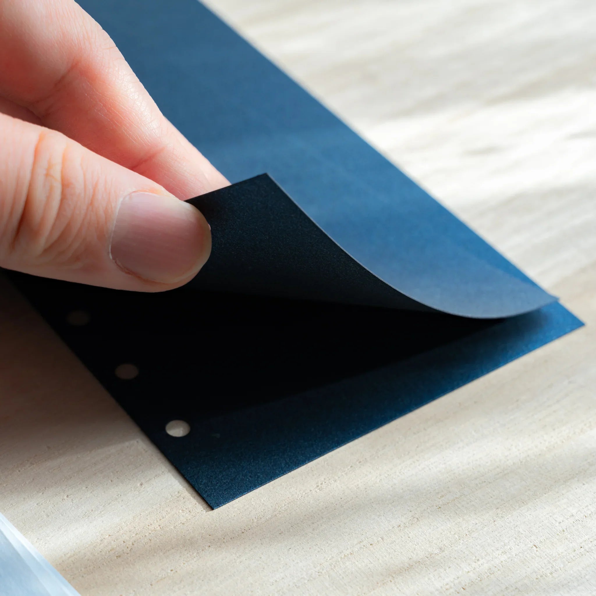 Hand lifting a dark navy onion skin planner sheet, showing its thin texture and six-hole punches on a light desk.