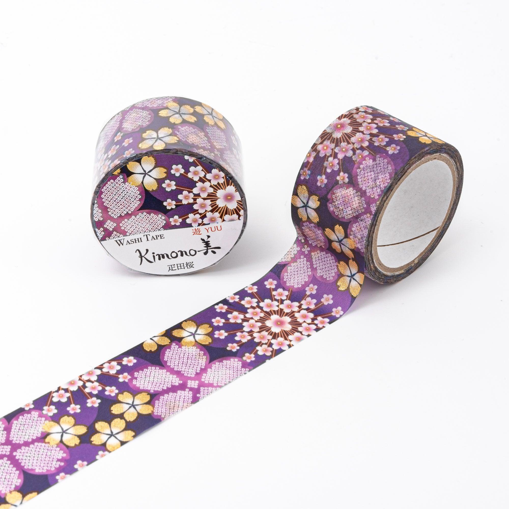 Unrolled Japanese stationery washi tape highlighting detailed floral motifs, including sakura, in pink and gold tones on a rich purple surface.
