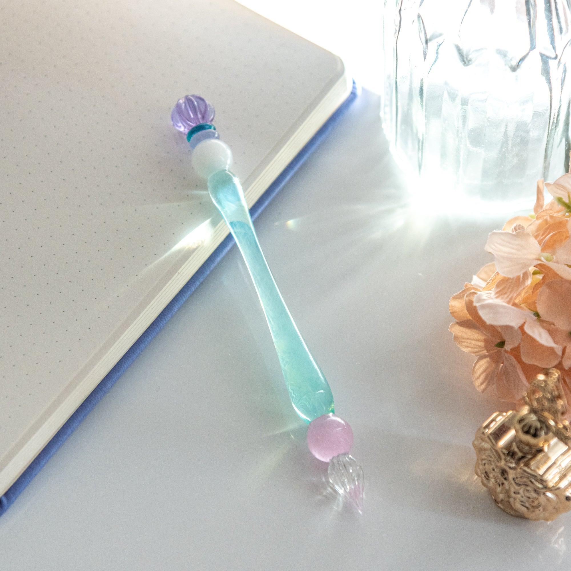 A glass pen with pastel accents, featuring a purple candy-like ornament and a soft blue body with a pink bead near the nib.
