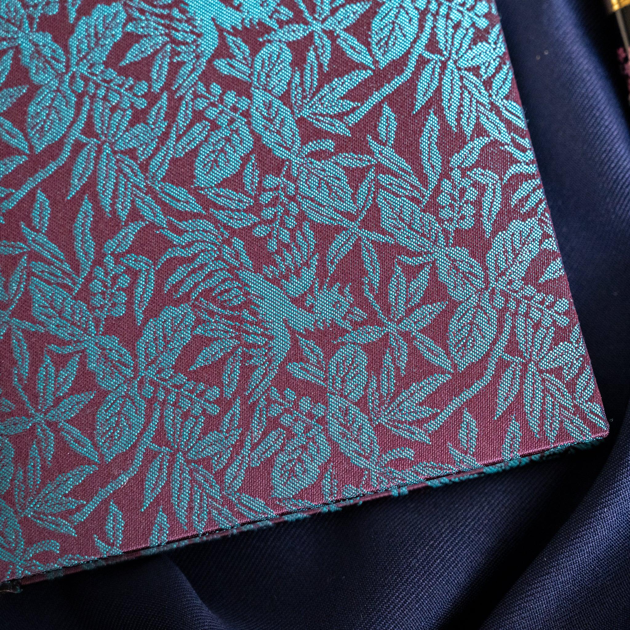 Close-up detail of Kiri Take Ho-o Nishijin silk notebook cover showing teal leaf and bamboo woven pattern on maroon