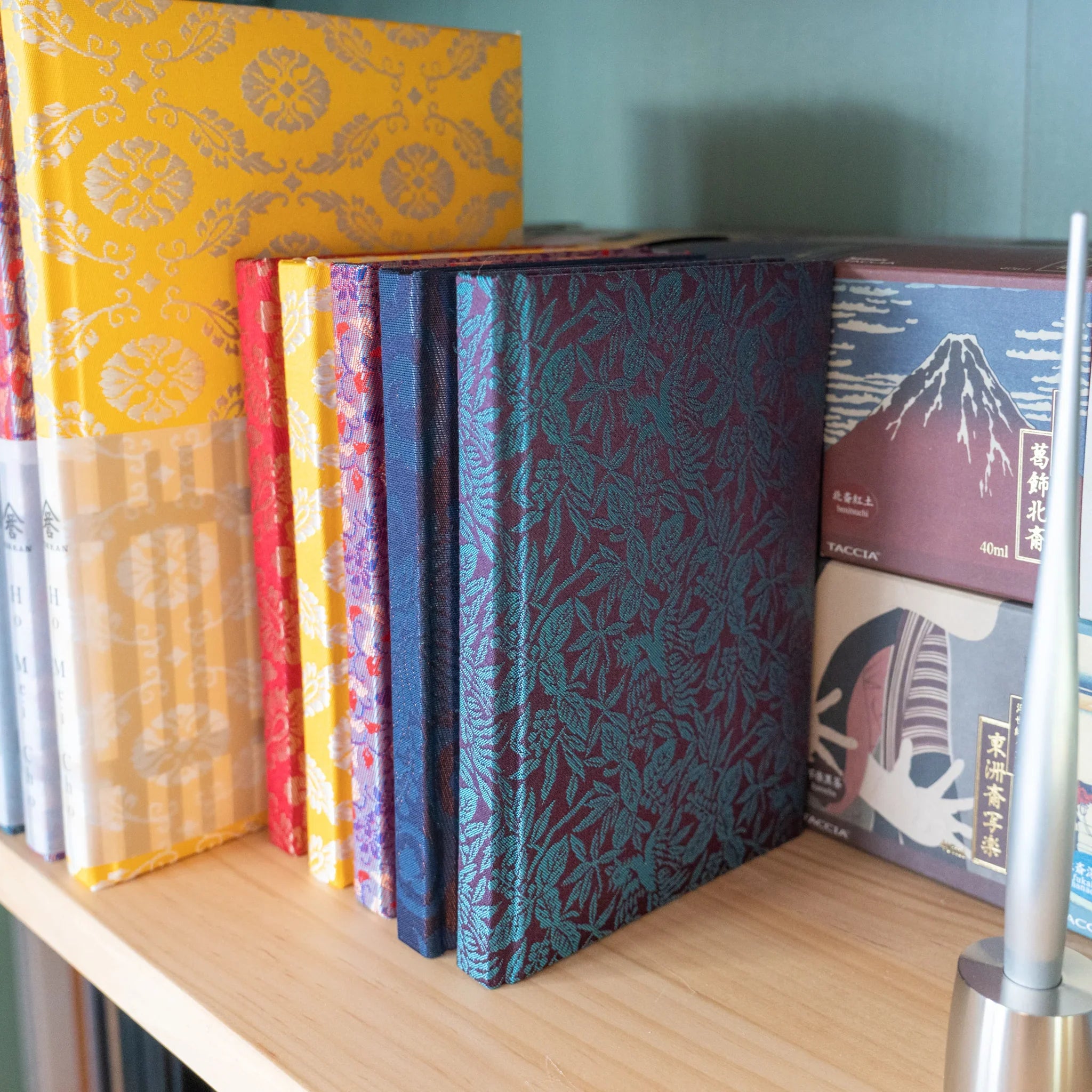 Multiple Nishijin silk Homeicho notebooks on wooden shelf showing Kiri Take Ho-o teal-on-maroon cover at front