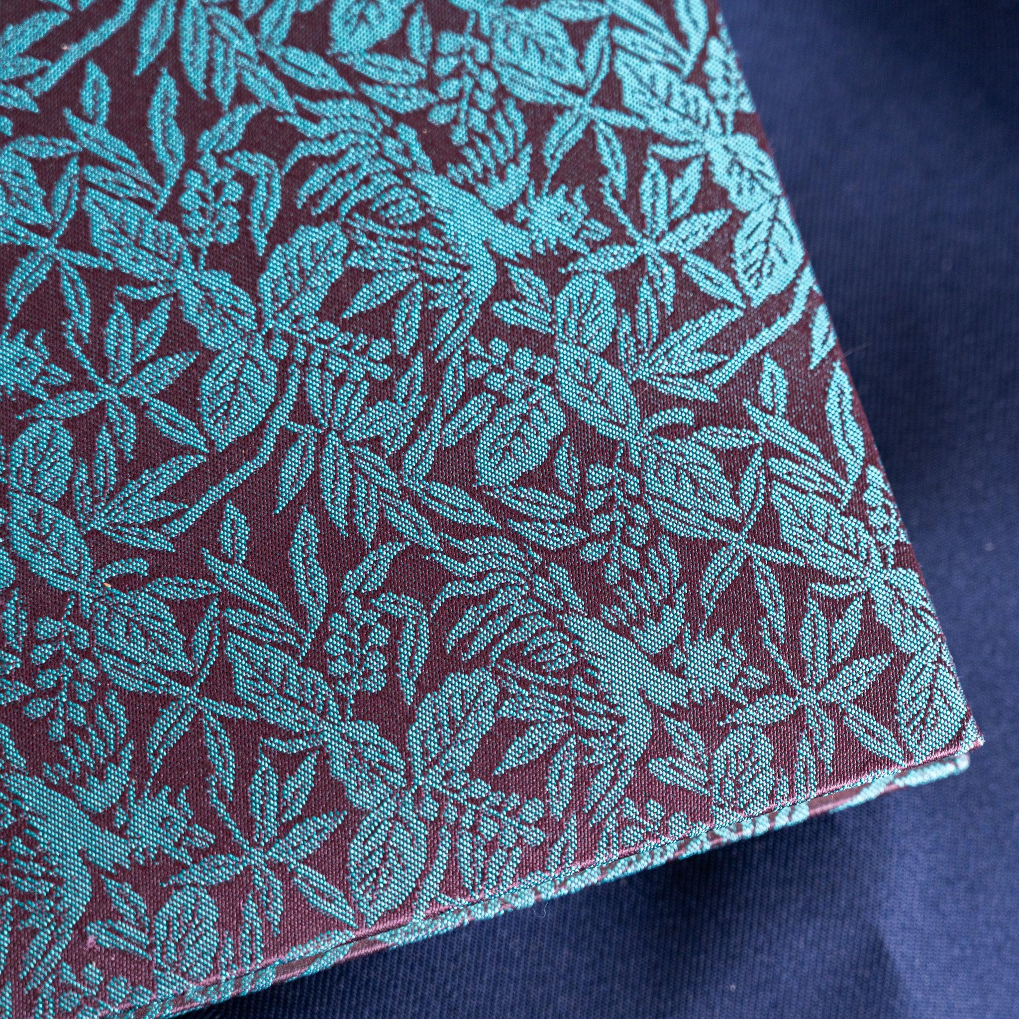 Macro close-up of Kiri Take Ho-o Nishijin silk notebook cover corner showing detailed teal leaf weave on maroon ground