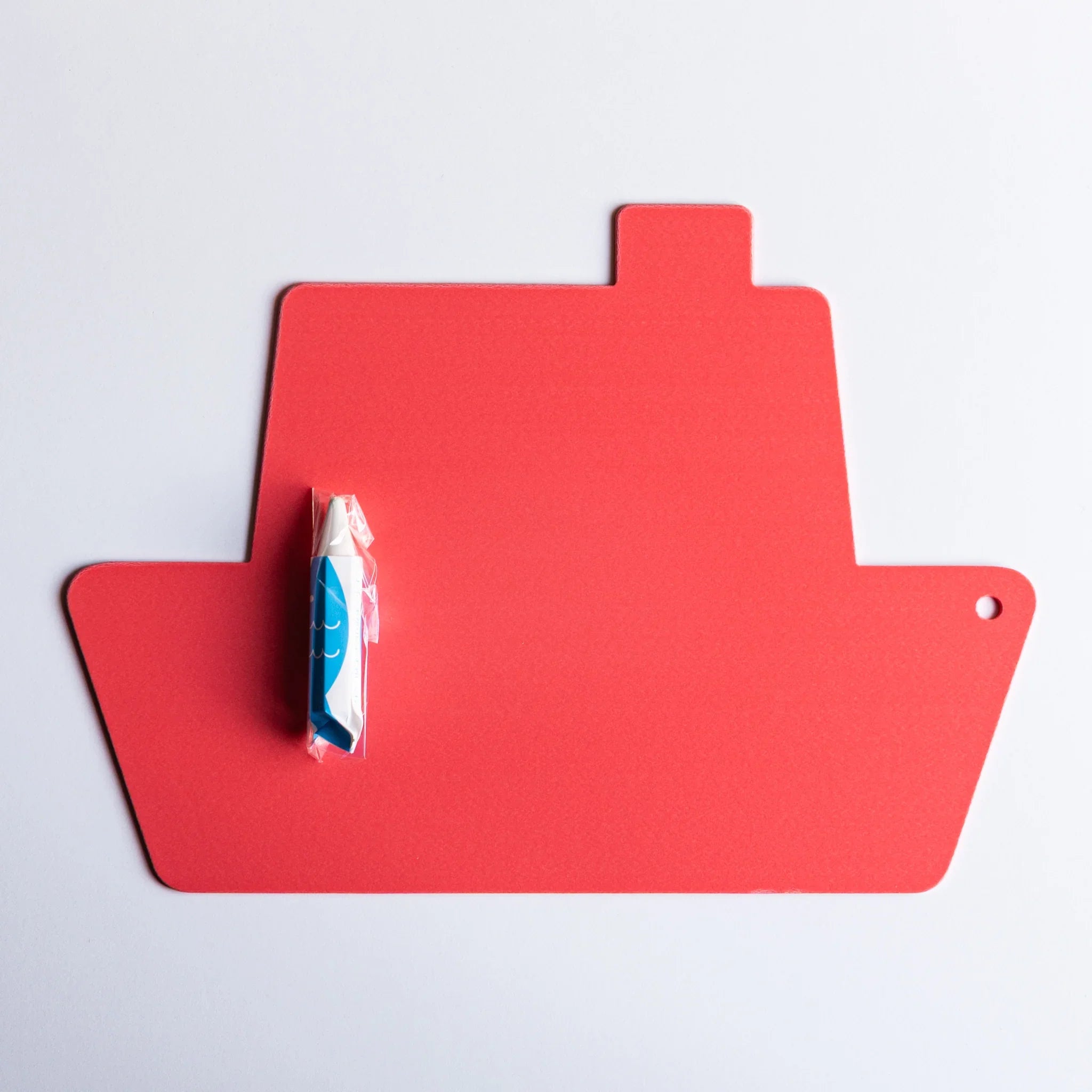 Red ship-shaped kitpas bath drawing board with a blue and white kitpas crayon sealed in plastic on a white surface.