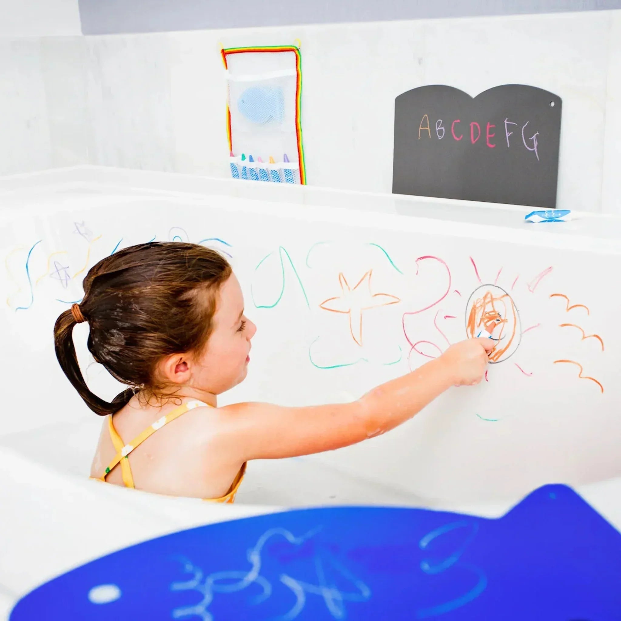 Child uses kitpas watercolor crayons to draw colorful stars, fish and sun designs on a white bathtub wall during bath time