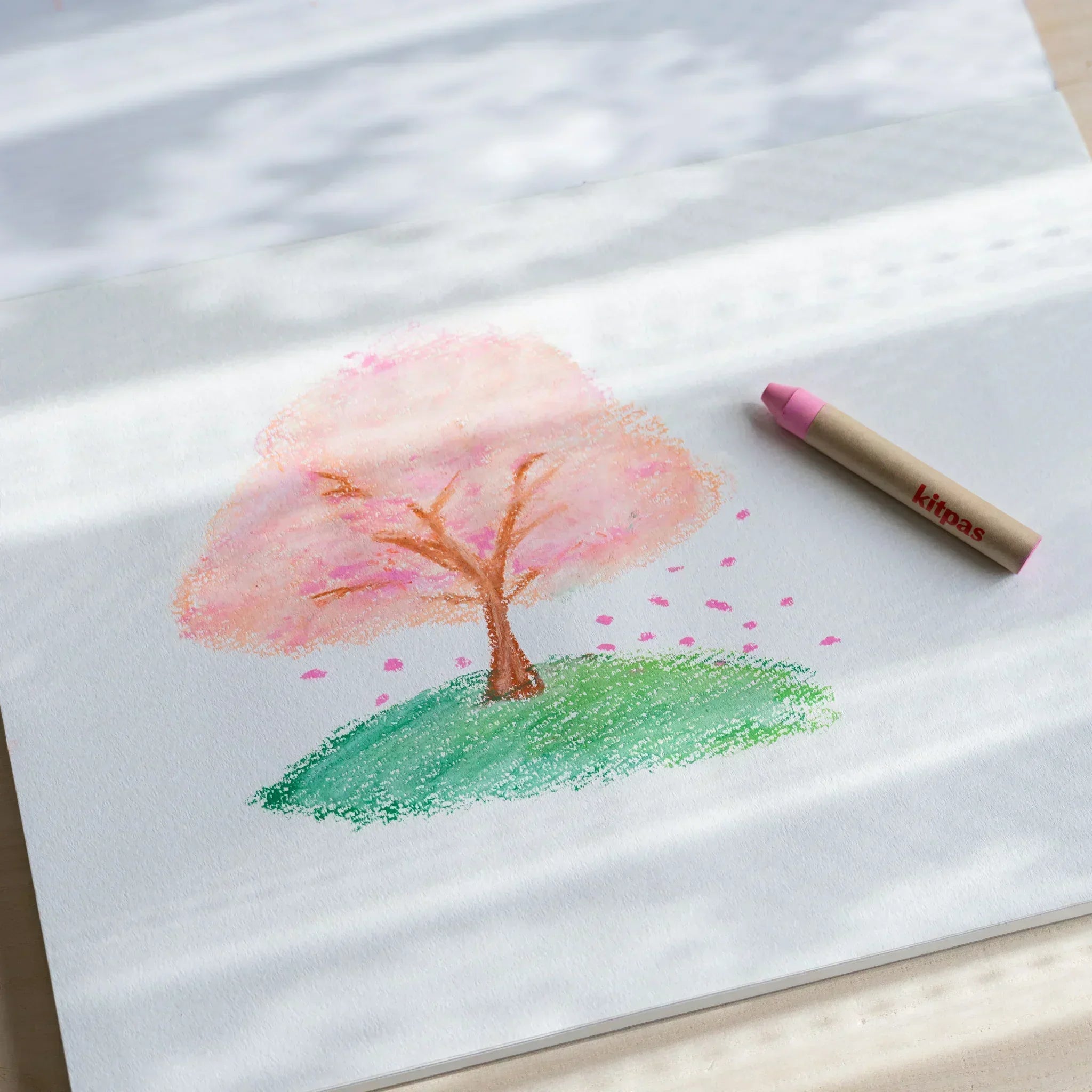 Cherry blossom tree drawing made with pink and green kitpas watercolor crayons on white paper, with a single kitpas crayon beside it.