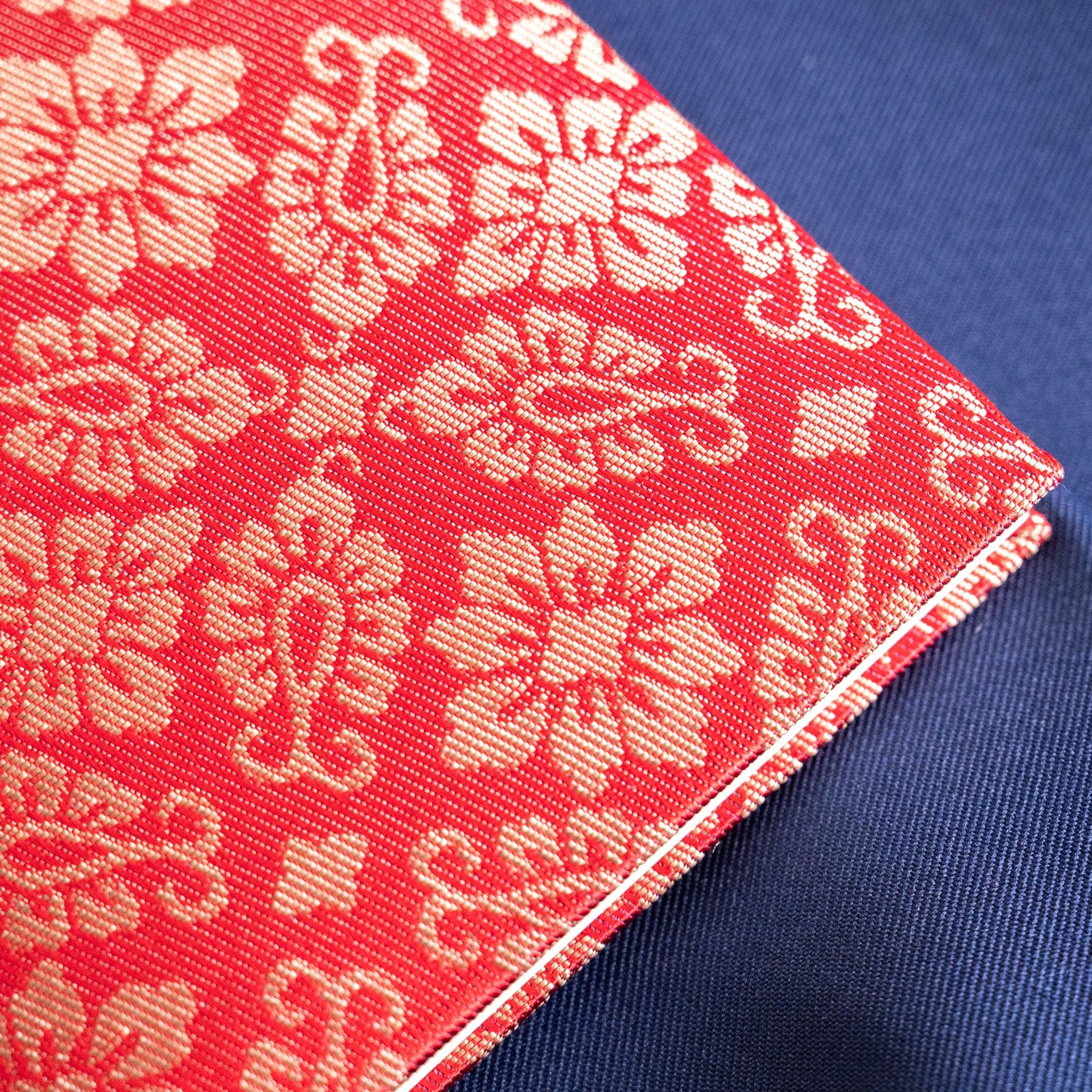 Angled corner close-up of Ko Aoi Nishijin silk cover showing vermilion hollyhock weave pattern and fabric binding edge