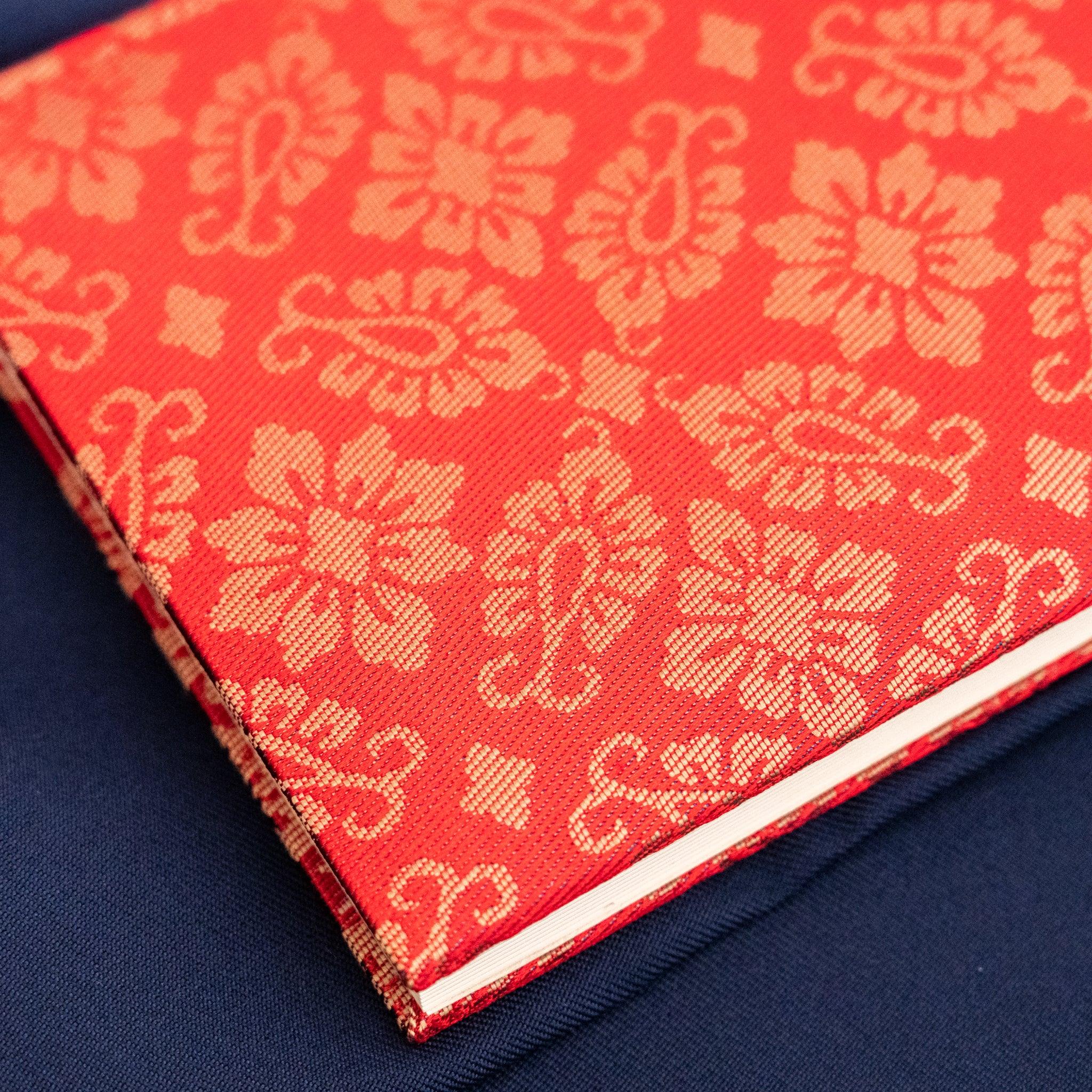 Ko Aoi Nishijin silk Homeicho notebook bottom corner showing vermilion hollyhock cover and white page edges on blue fabric