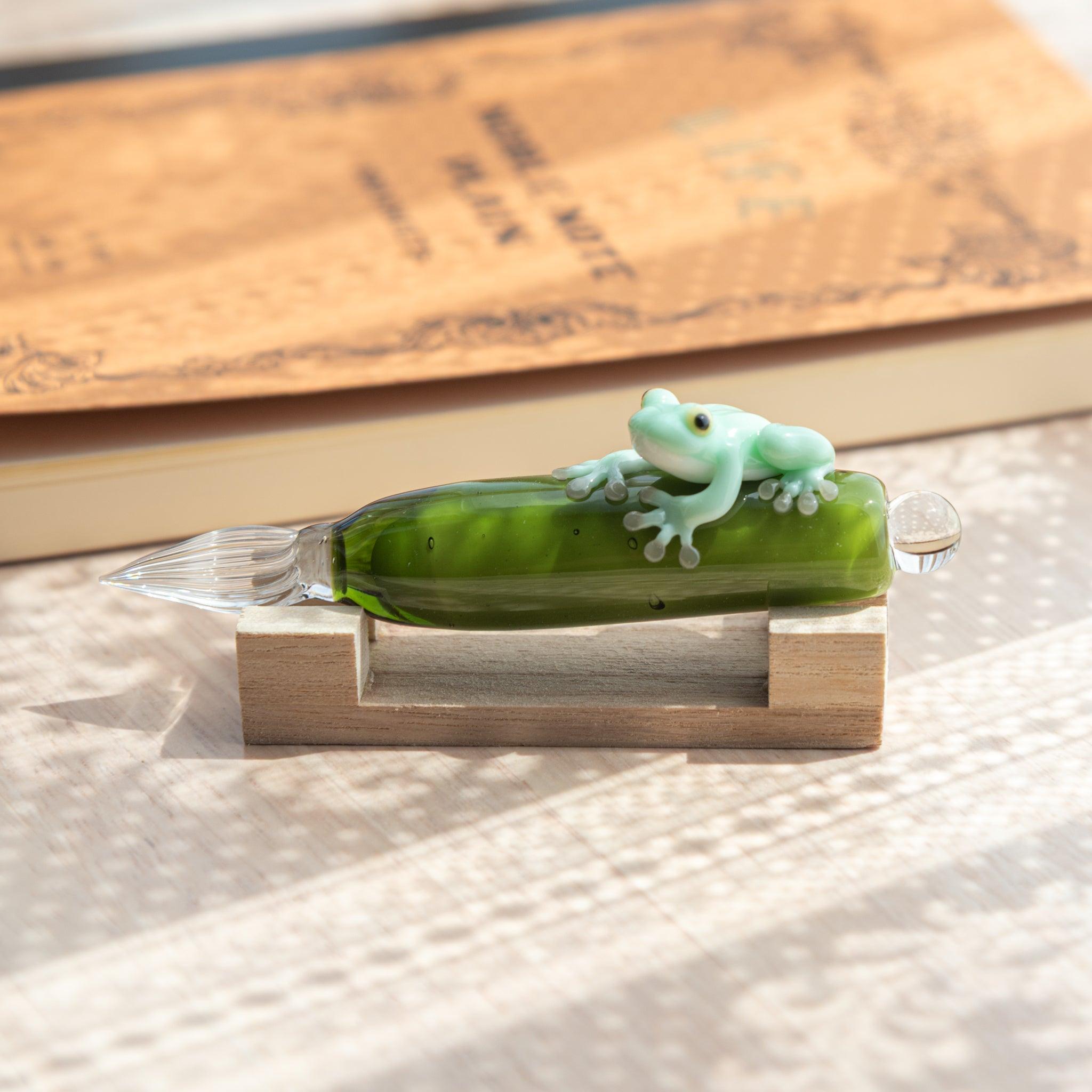 Komorebi Frog Glass Dip Pen on a wooden pen rest, featuring a light green frog and clear glass nib under soft natural light.