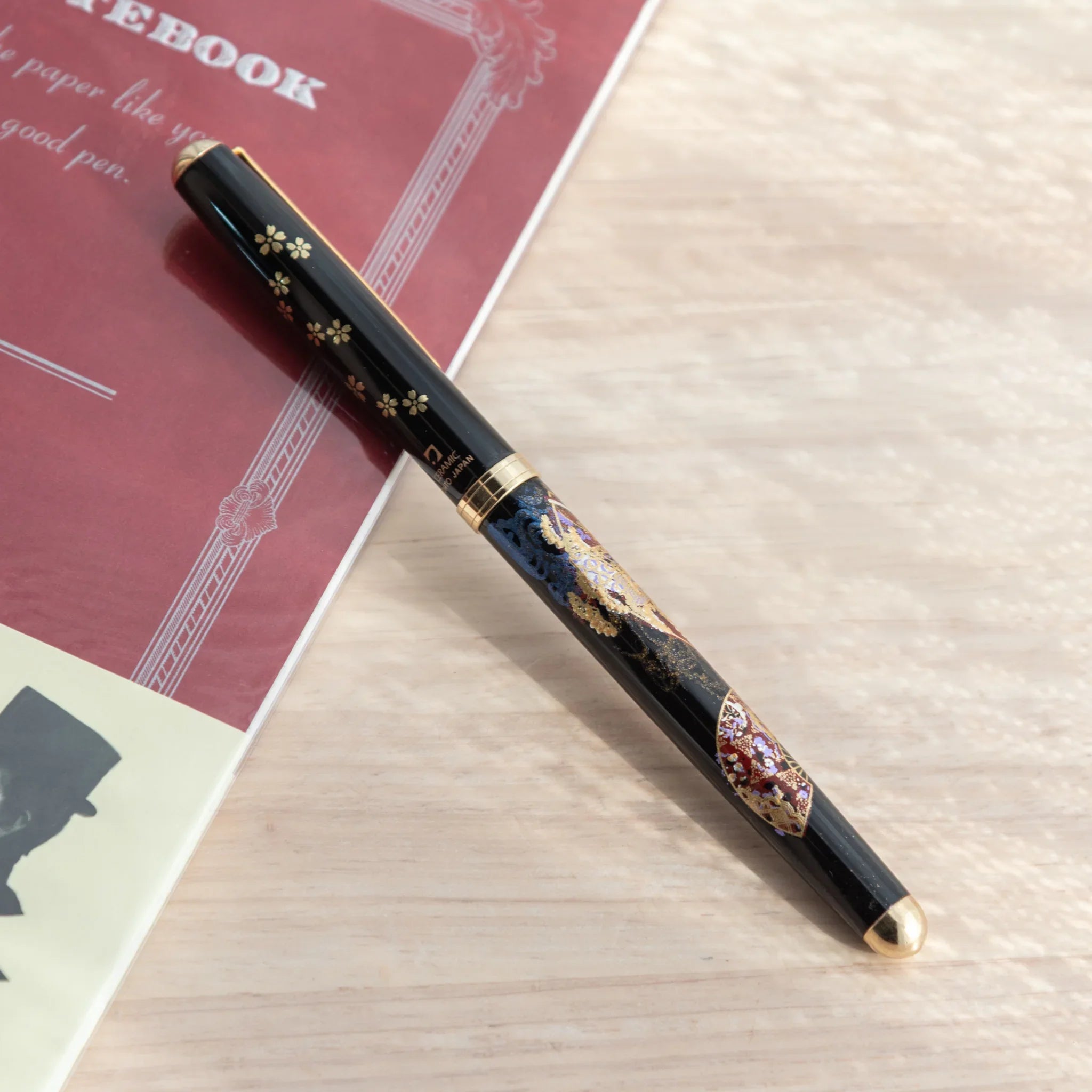Black Yamanaka lacquer Japanese ballpoint pen with Kyoto fan and maiko maki-e barrel design and sakura cap, near red notebook