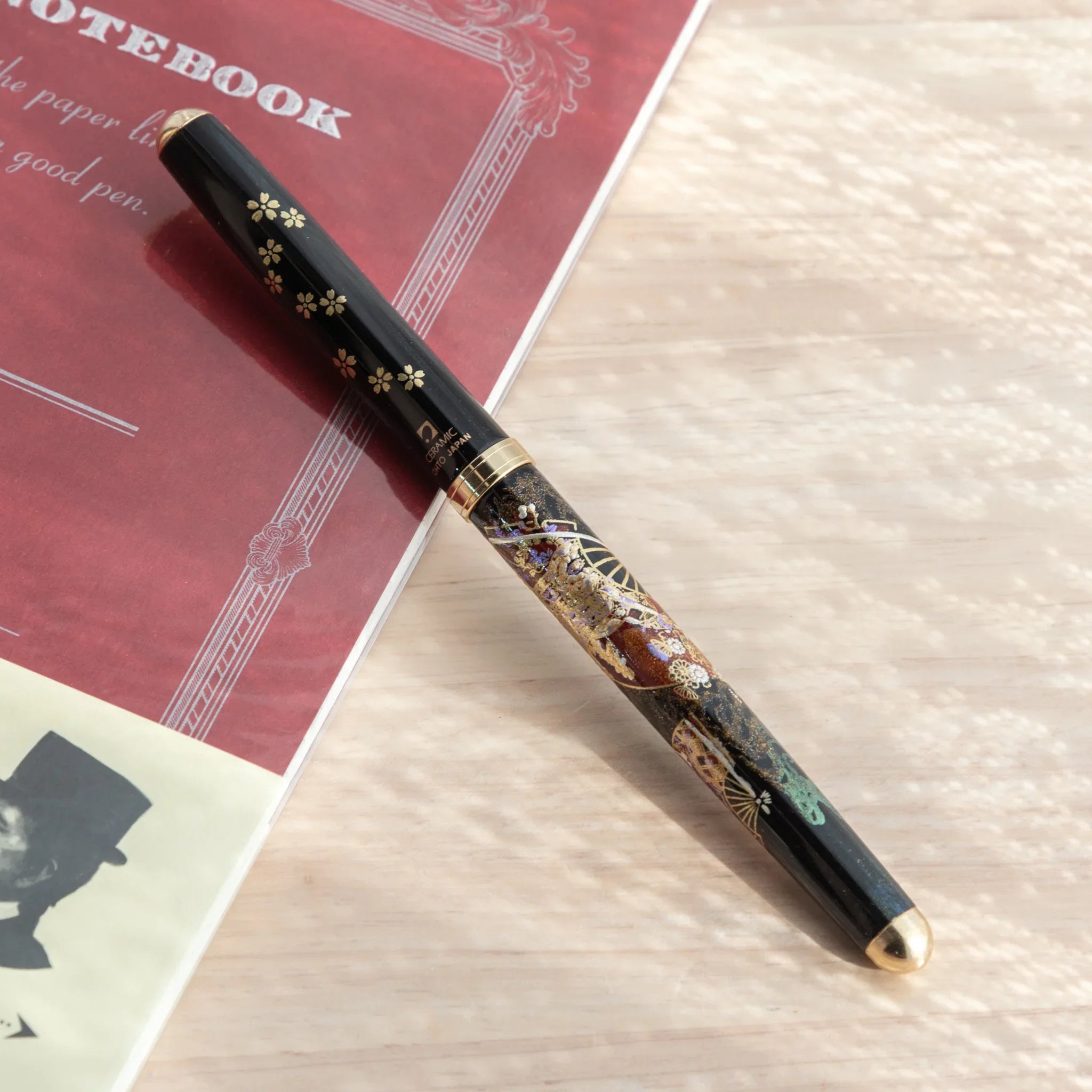 Yamanaka Japanese ballpoint pen rotated to show teal pine, fan, and gold lacquer maki-e motifs on black barrel near notebook