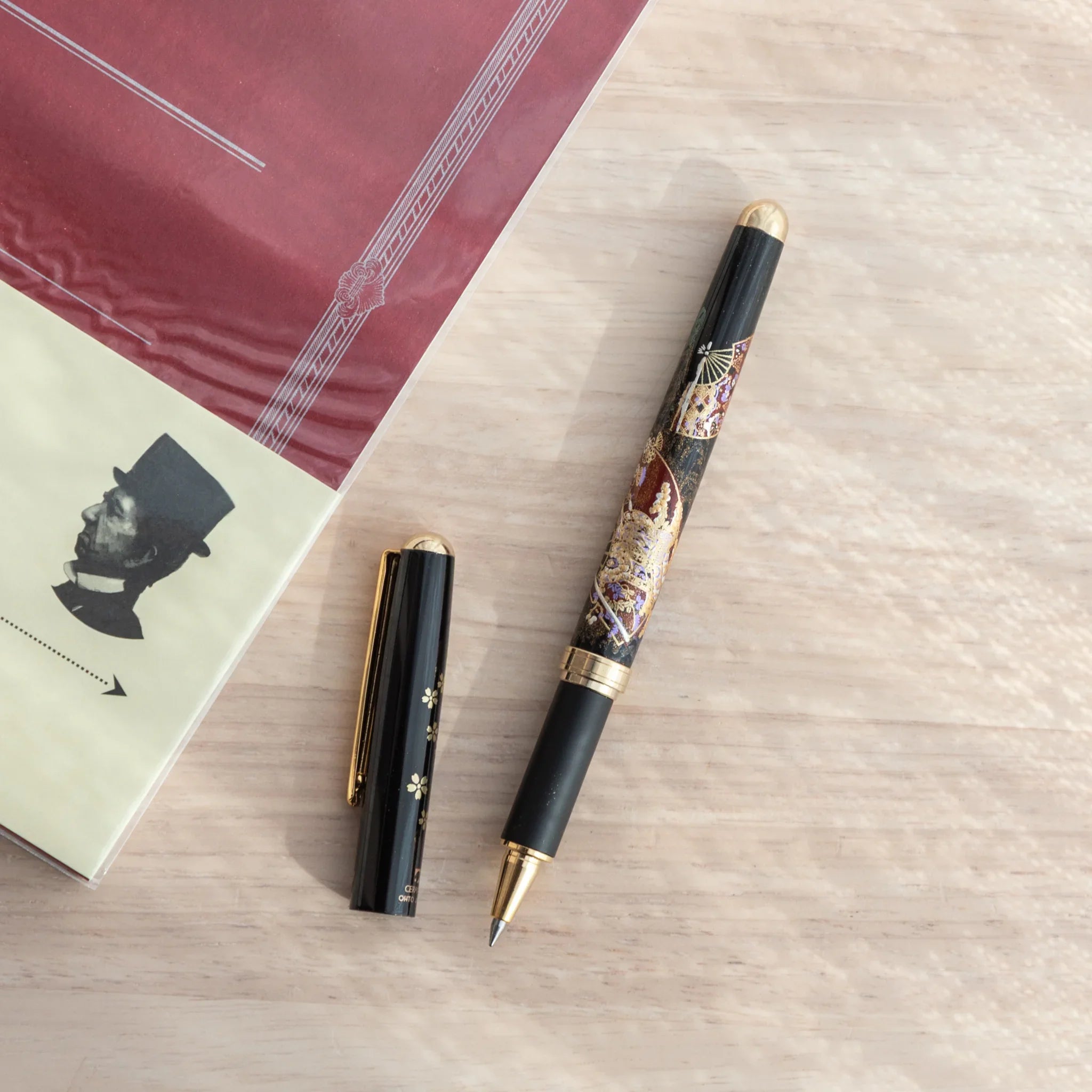 Japanese Ballpoint Pen with Lacquer artwork of cherry blossoms and fan, placed on red C.D. notebook under natural light.