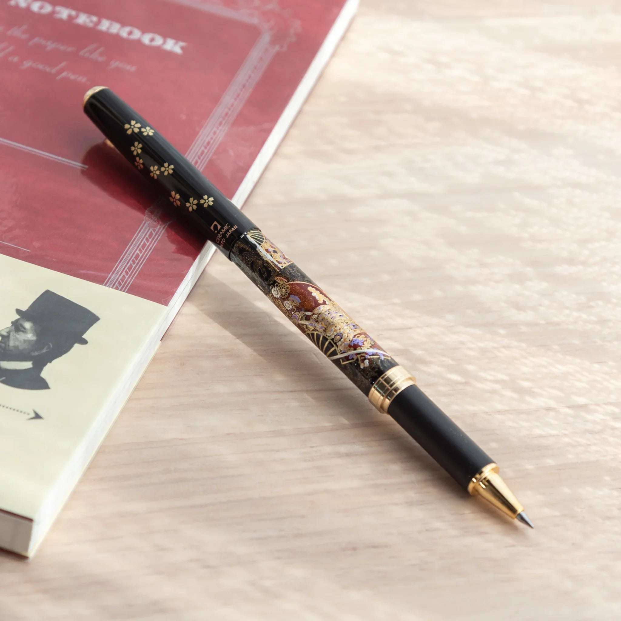 Yamanaka Japanese ballpoint pen uncapped with tip forward, black barrel showing fan and maiko lacquer design, on red notebook
