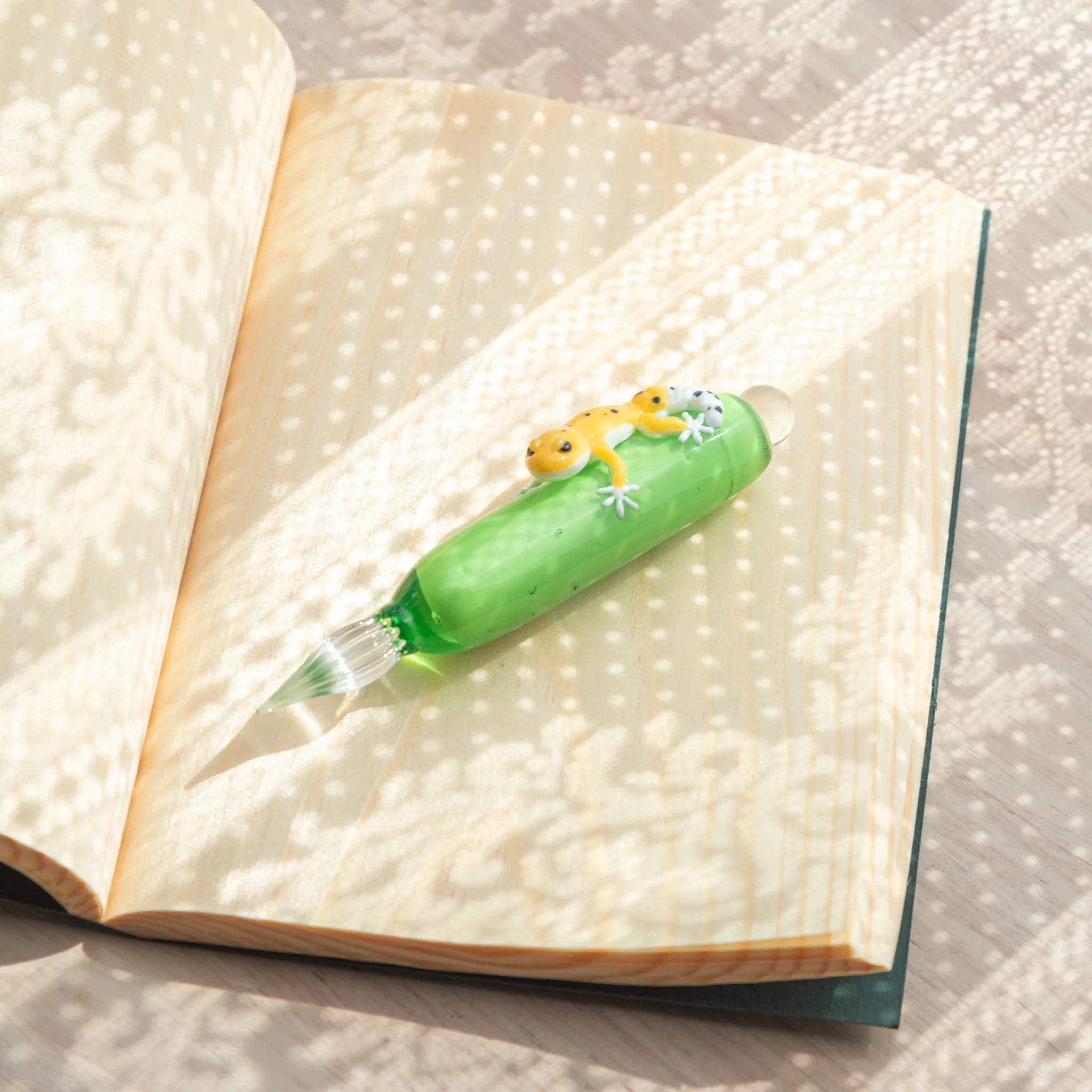 Green glass dip pen with yellow spotted leopard gecko on barrel lying on open cream dot-grid notebook pages