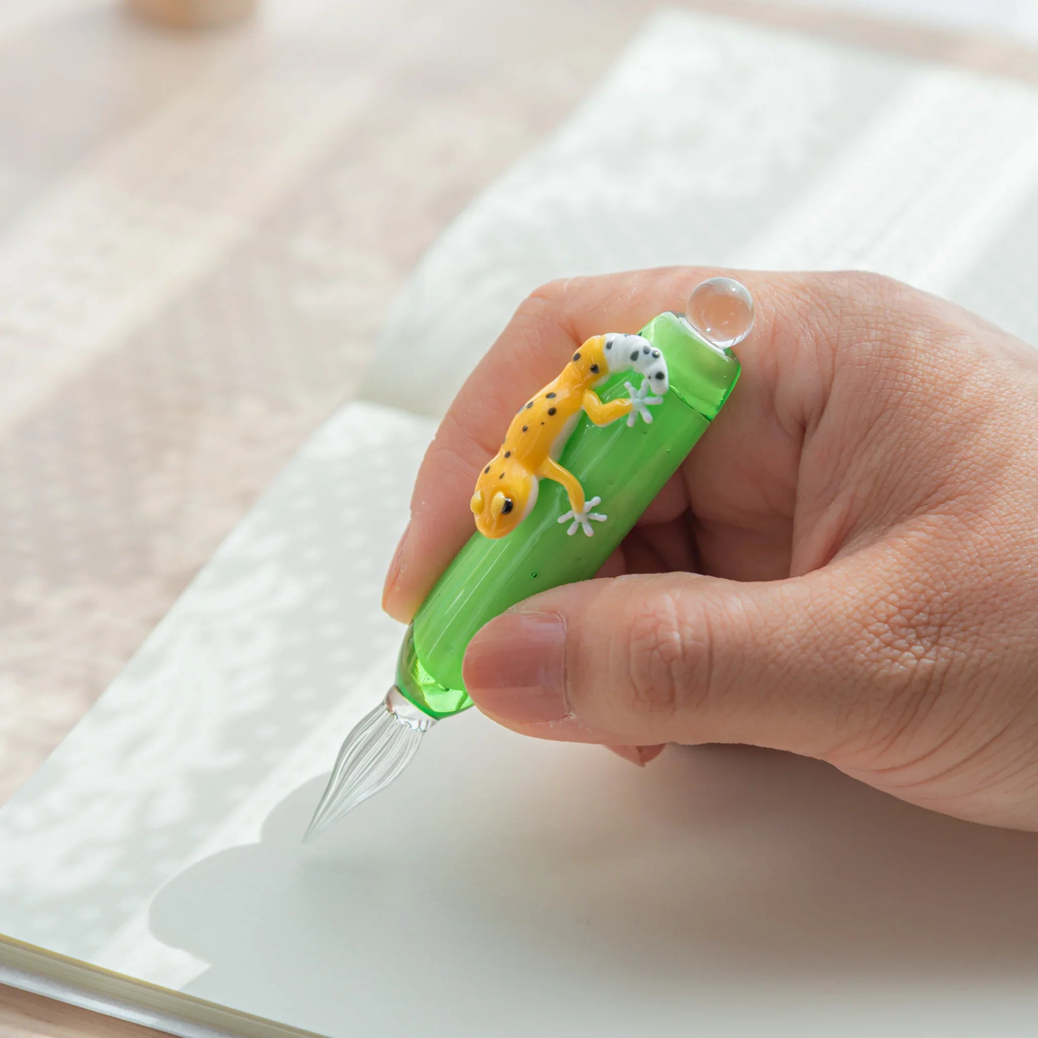 Hand holding green glass dip pen over open notebook showing yellow spotted leopard gecko on barrel and ribbed glass nib