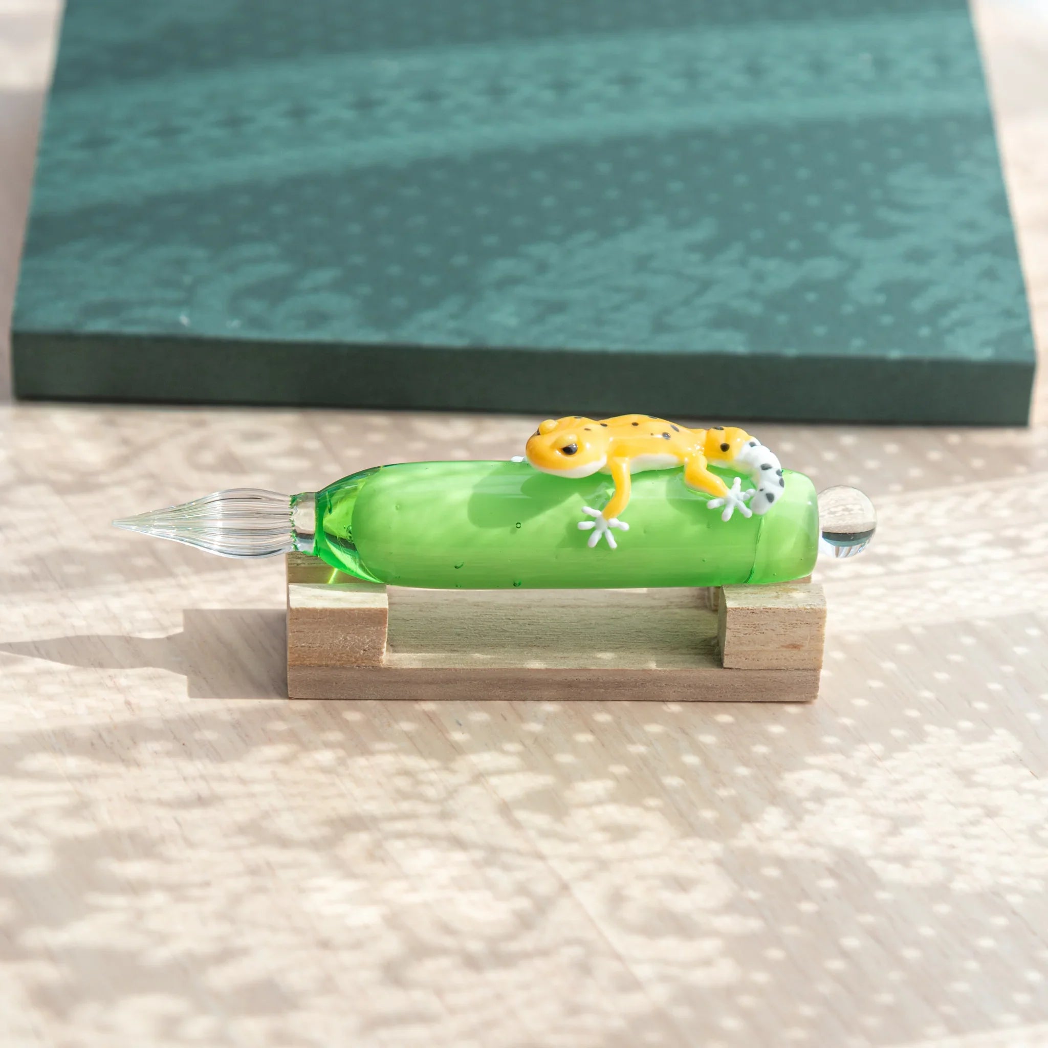 Green glass dip pen on wood pen stand showing yellow spotted leopard gecko on barrel and glass nib, teal notebook behind