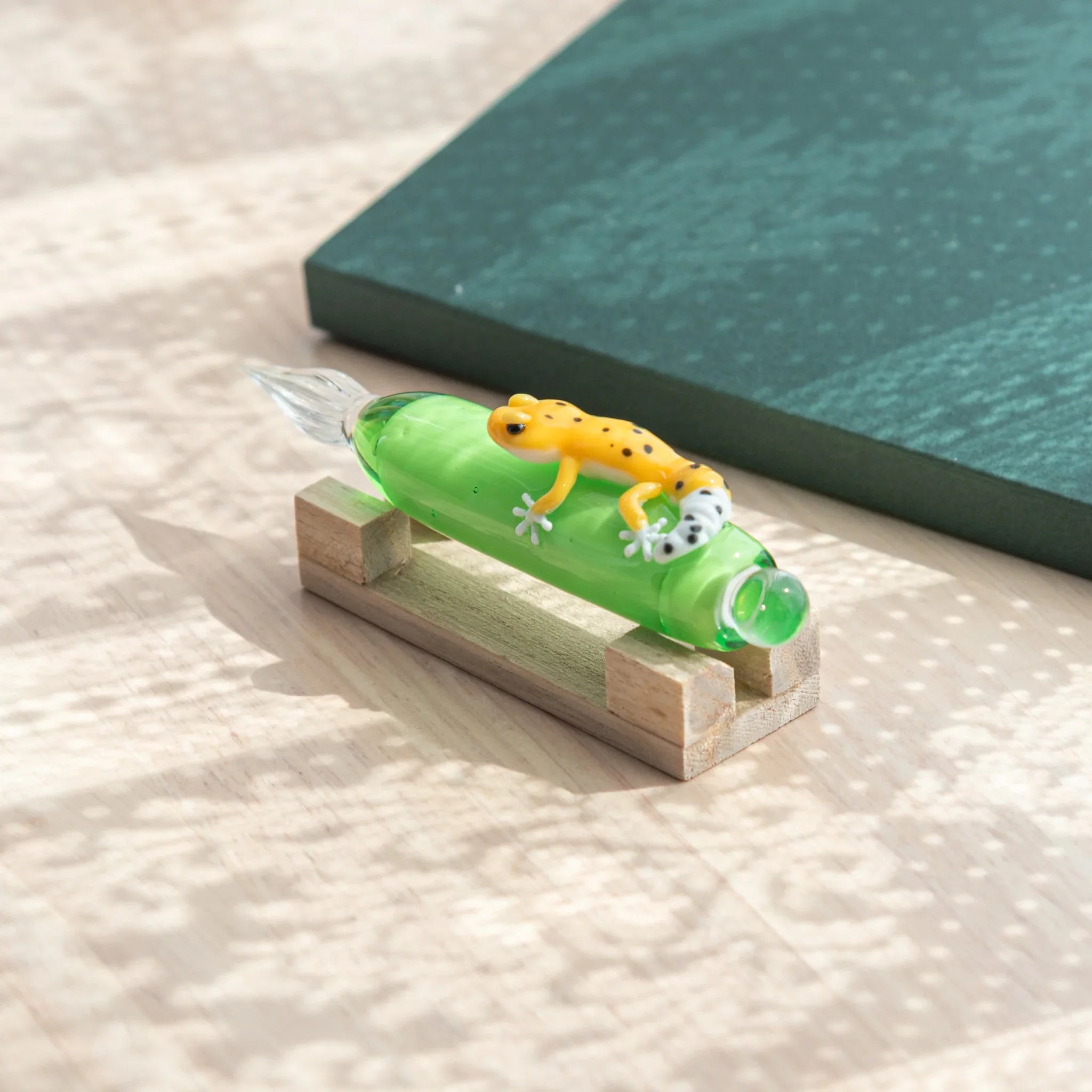 Green glass dip pen on wood pen stand, front-angled view, showing yellow spotted leopard gecko figure on barrel and glass nib