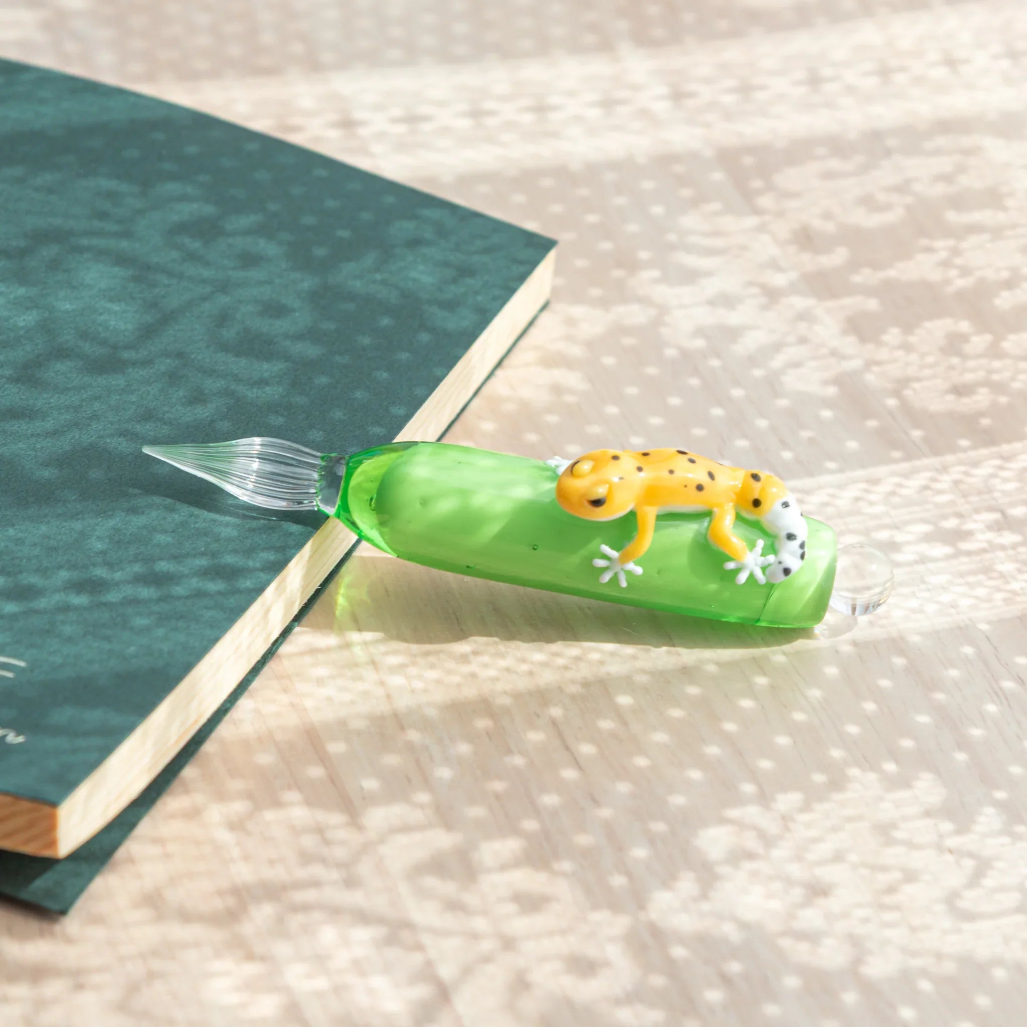 Green glass dip pen on desk beside teal notebook with yellow spotted leopard gecko figure on barrel and clear glass nib