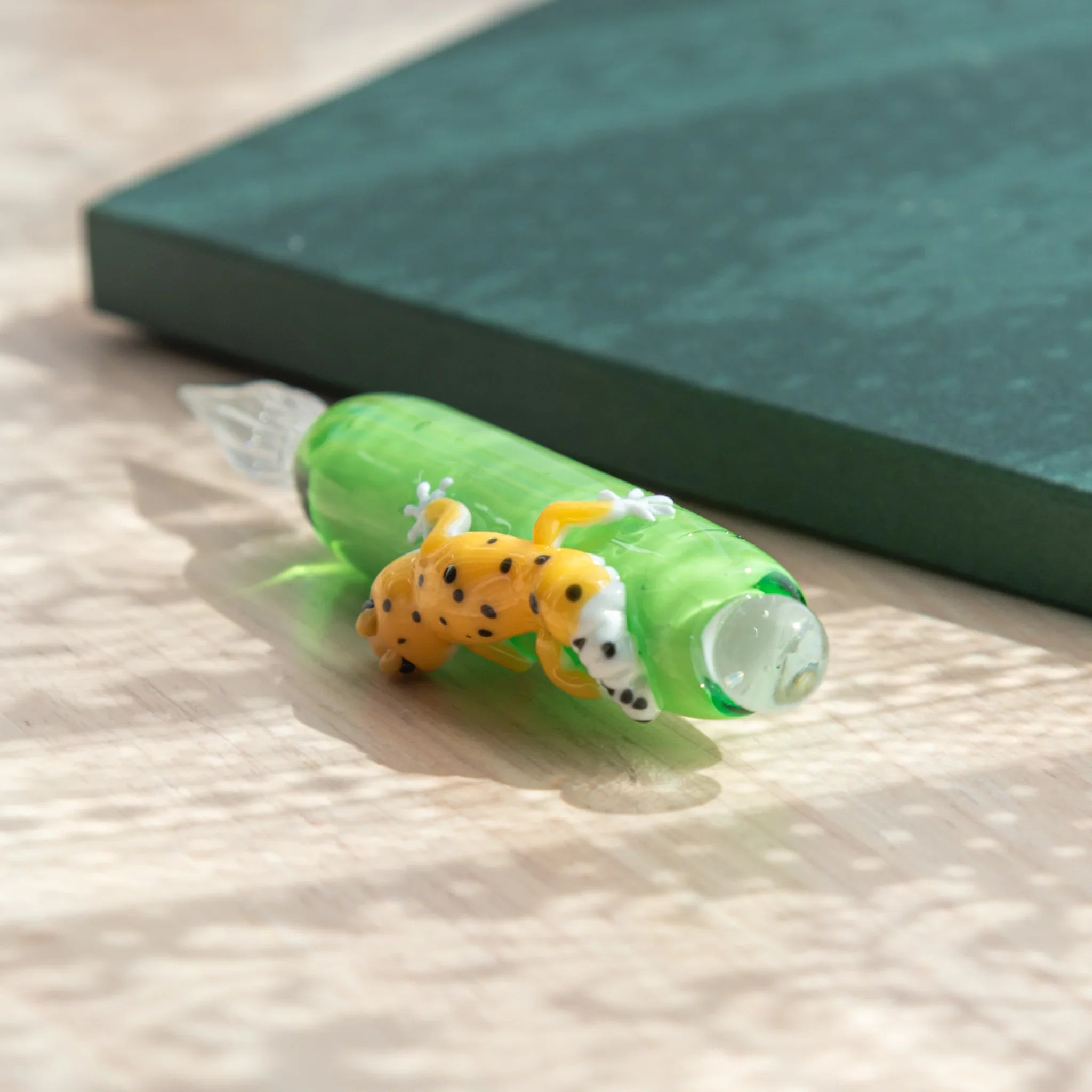 Green glass dip pen viewed from above beside teal notebook, showing yellow black-spotted leopard gecko figure on barrel