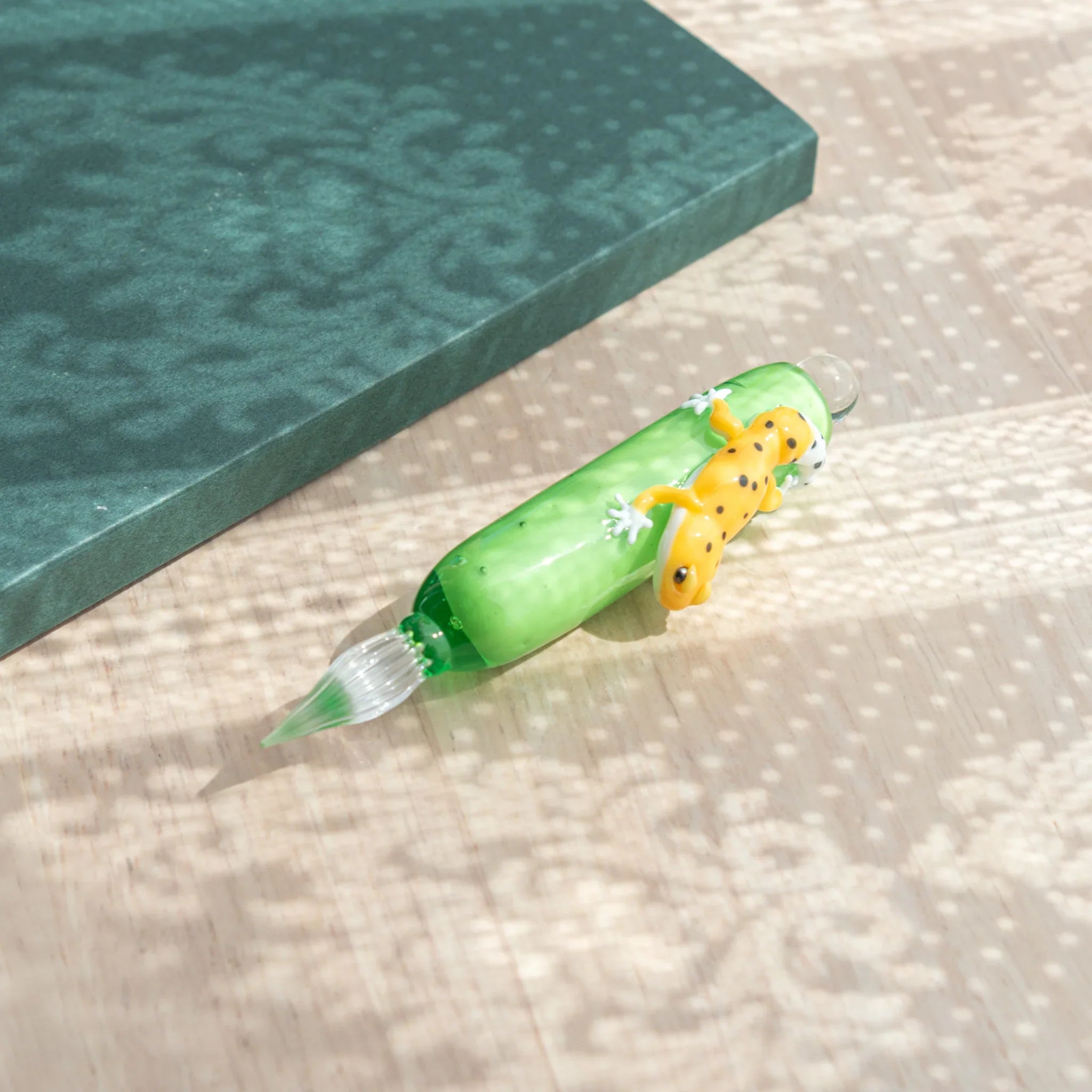 Green glass dip pen on wood surface with nib angled down and yellow spotted leopard gecko on barrel, teal notebook behind