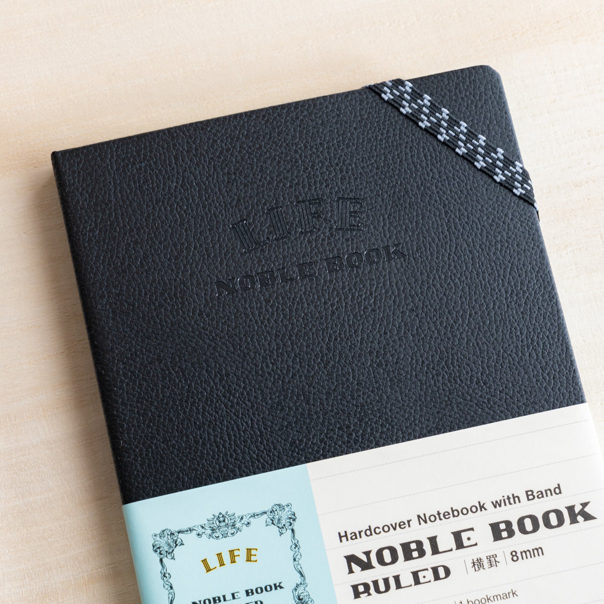 Life Noble notebook A6 hard cover front view with elastic corner band on desk, showing textured black cover and compact design
