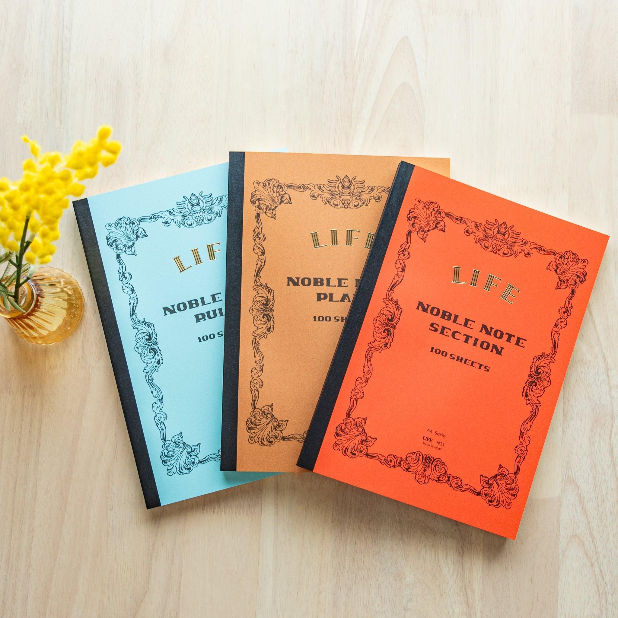 Three A4-size LIFE Noble Note notebooks (ruled, plain, grid) with pastel blue, brown, and orange covers placed on a wooden table.