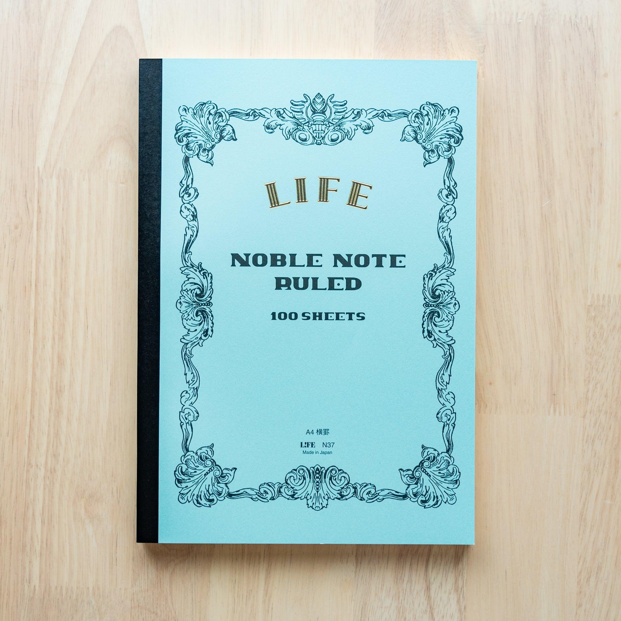 An A4-size LIFE Noble Note notebook with a pastel blue cover, gold lettering, and decorative black borders, labeled "Ruled, 100 Sheets."