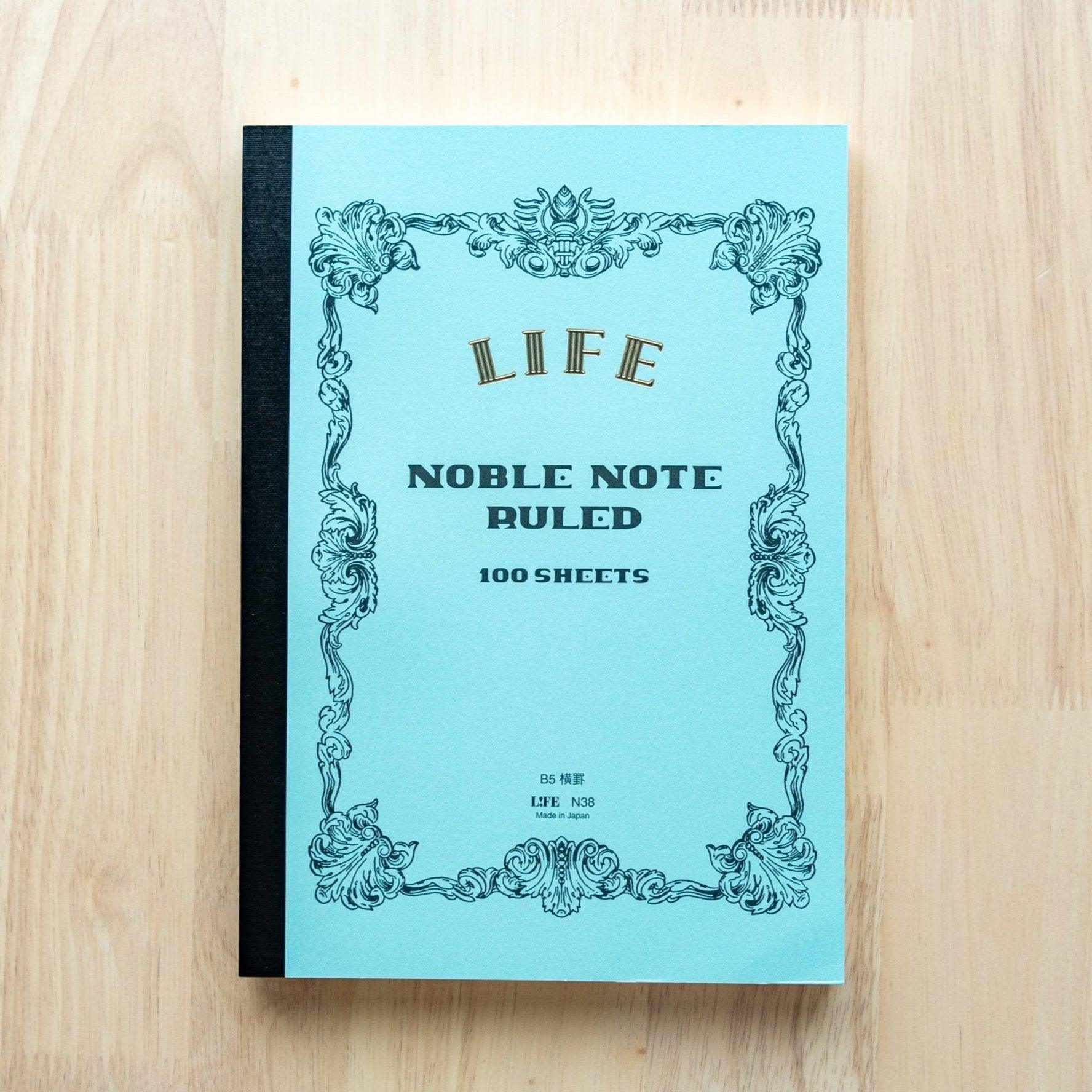 A single "LIFE Noble Note Ruled" notebook with a blue cover, black spine, ornate border design, and 100 sheets, displayed on a wooden surface.