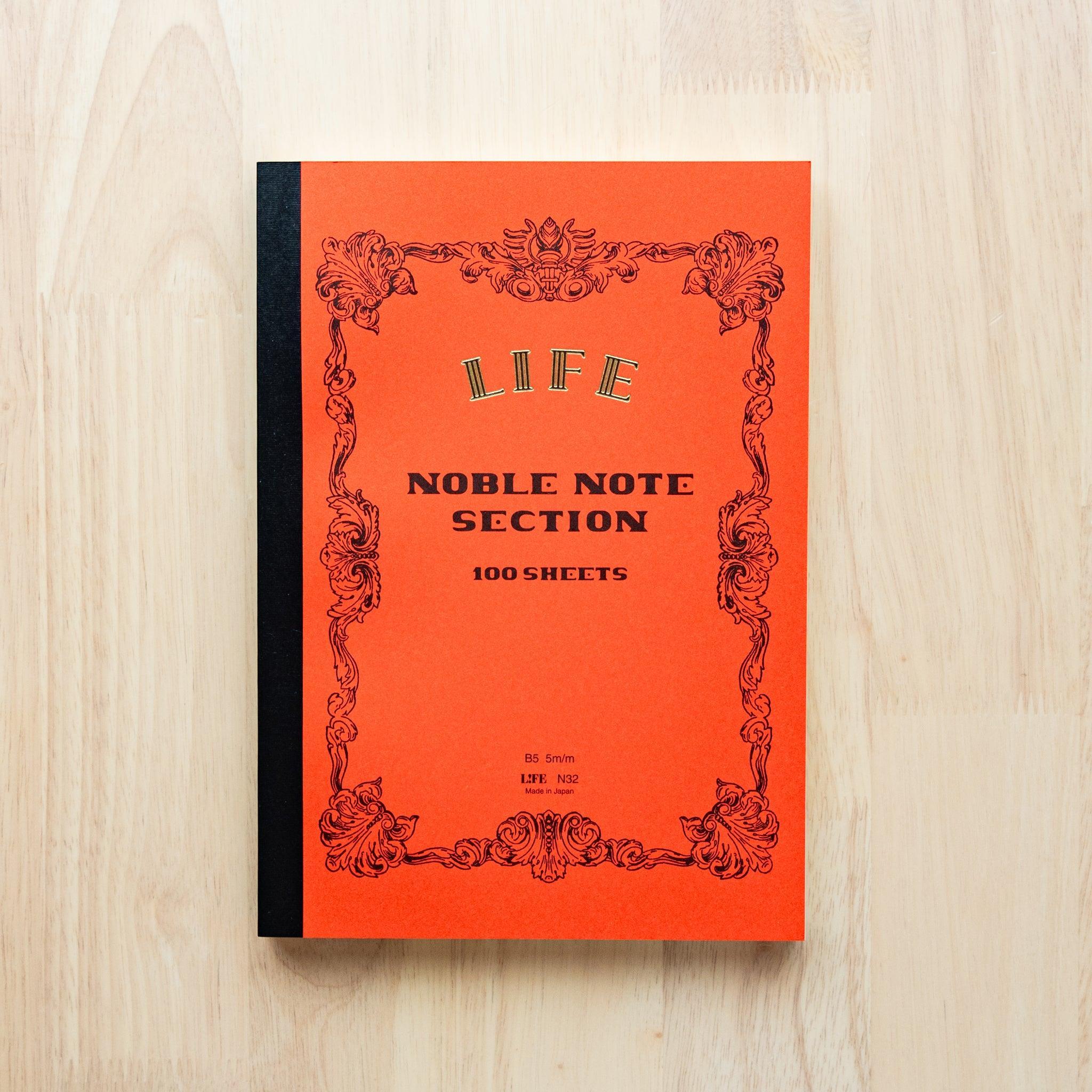 A single "LIFE Noble Note Ruled" notebook with a orange cover, black spine, ornate border design, and 100 sheets, displayed on a wooden surface.