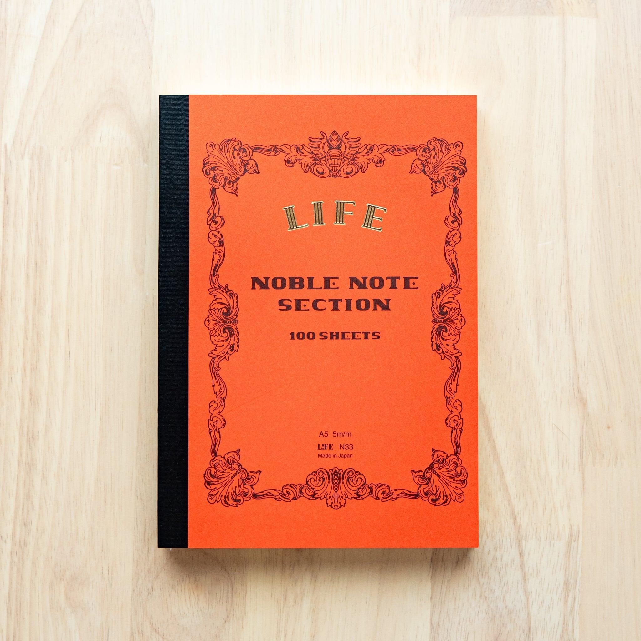 A single "LIFE Noble Note Ruled" notebook with a orange cover, black spine, ornate border design, and 100 sheets, displayed on a wooden surface.