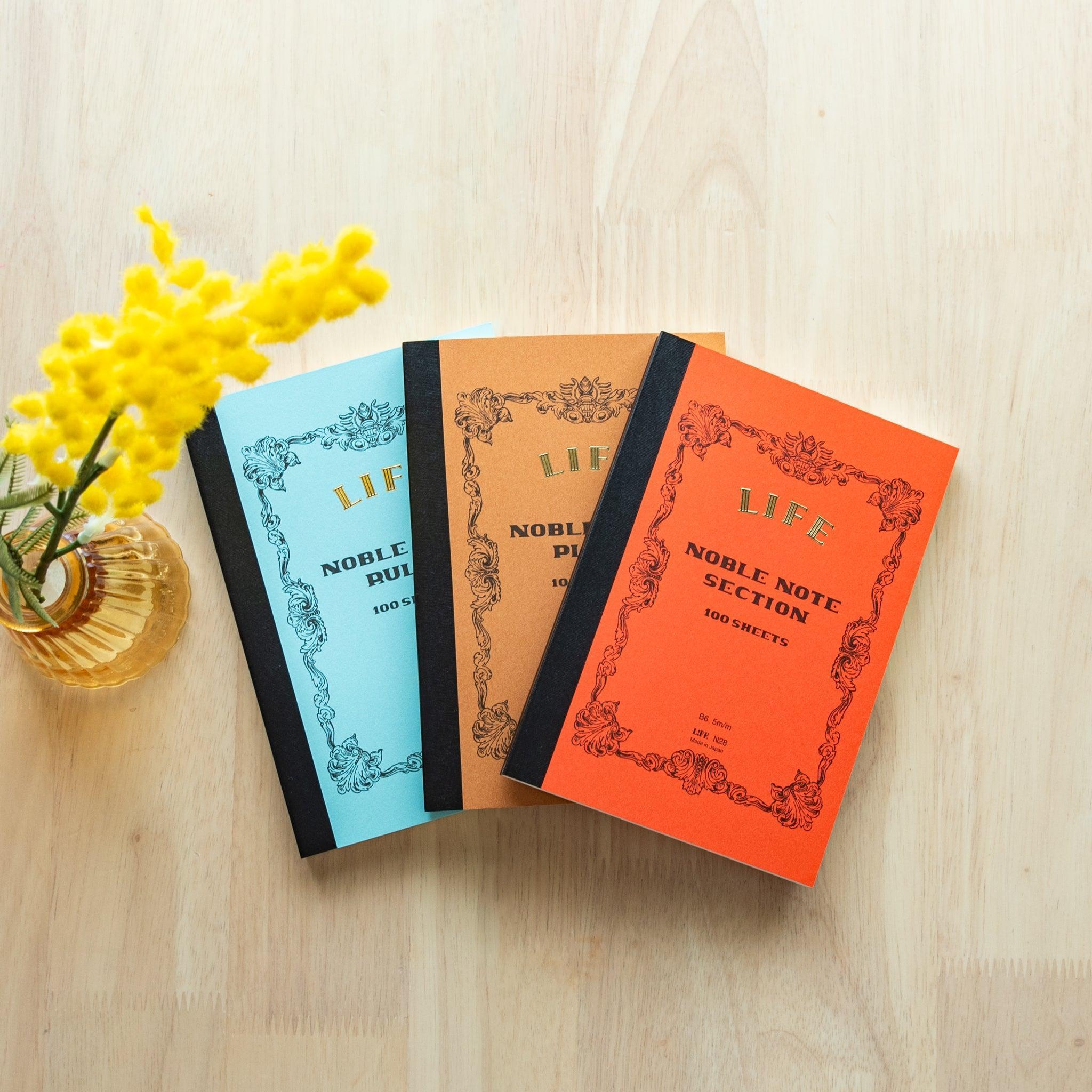 Three B6-size LIFE Noble Note notebooks (ruled, plain, grid) with pastel blue, brown, and orange covers placed on a wooden table.
