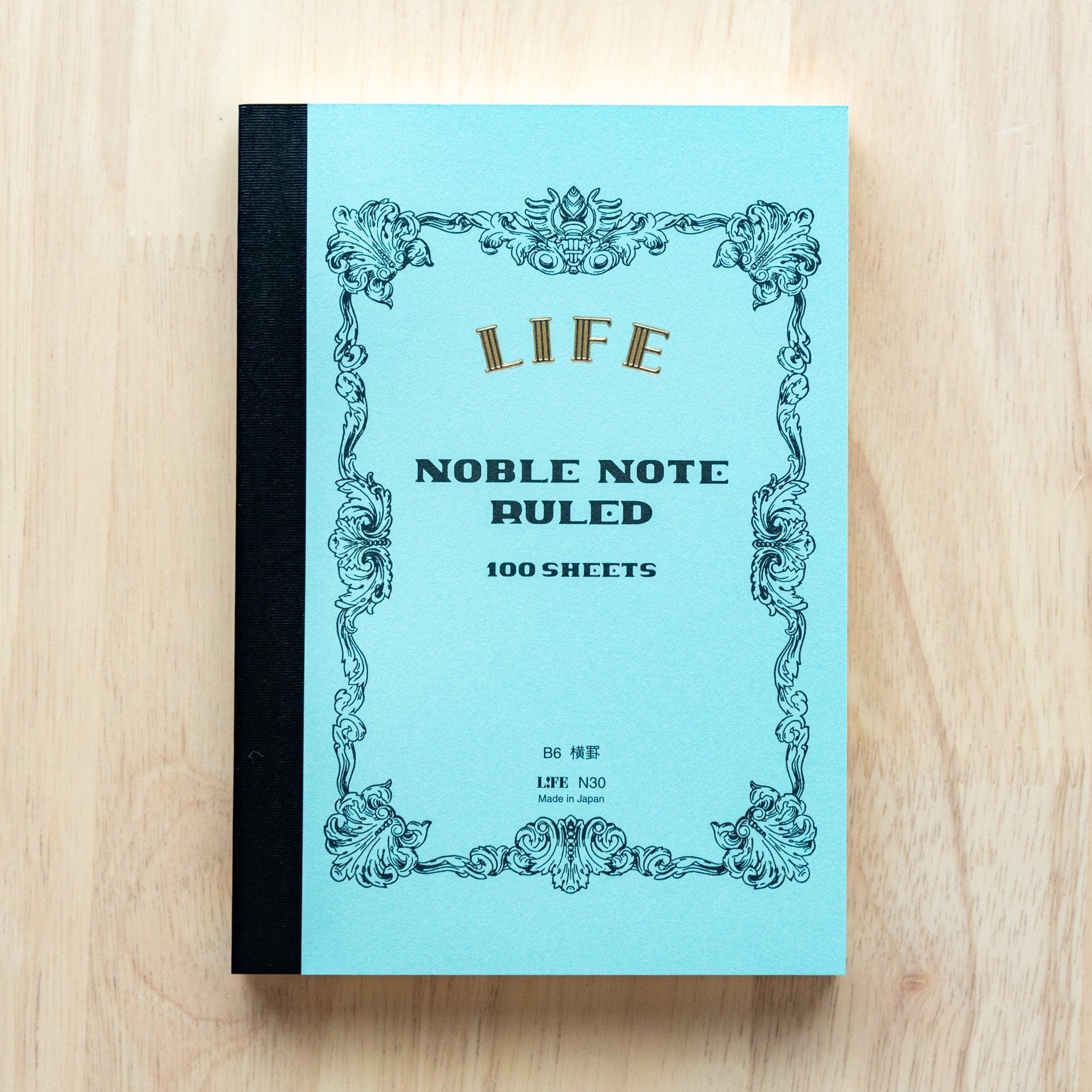 A single "LIFE Noble Note Ruled" notebook with a blue cover, black spine, ornate border design, and 100 sheets, displayed on a wooden surface.