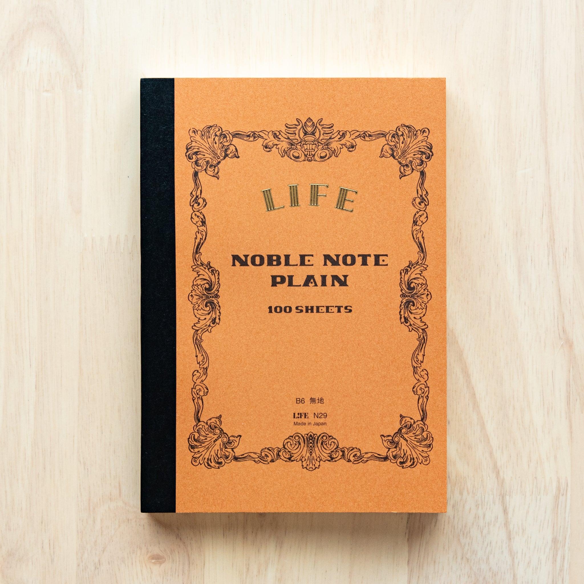 A single "LIFE Noble Note Ruled" notebook with a brown cover, black spine, ornate border design, and 100 sheets, displayed on a wooden surface.