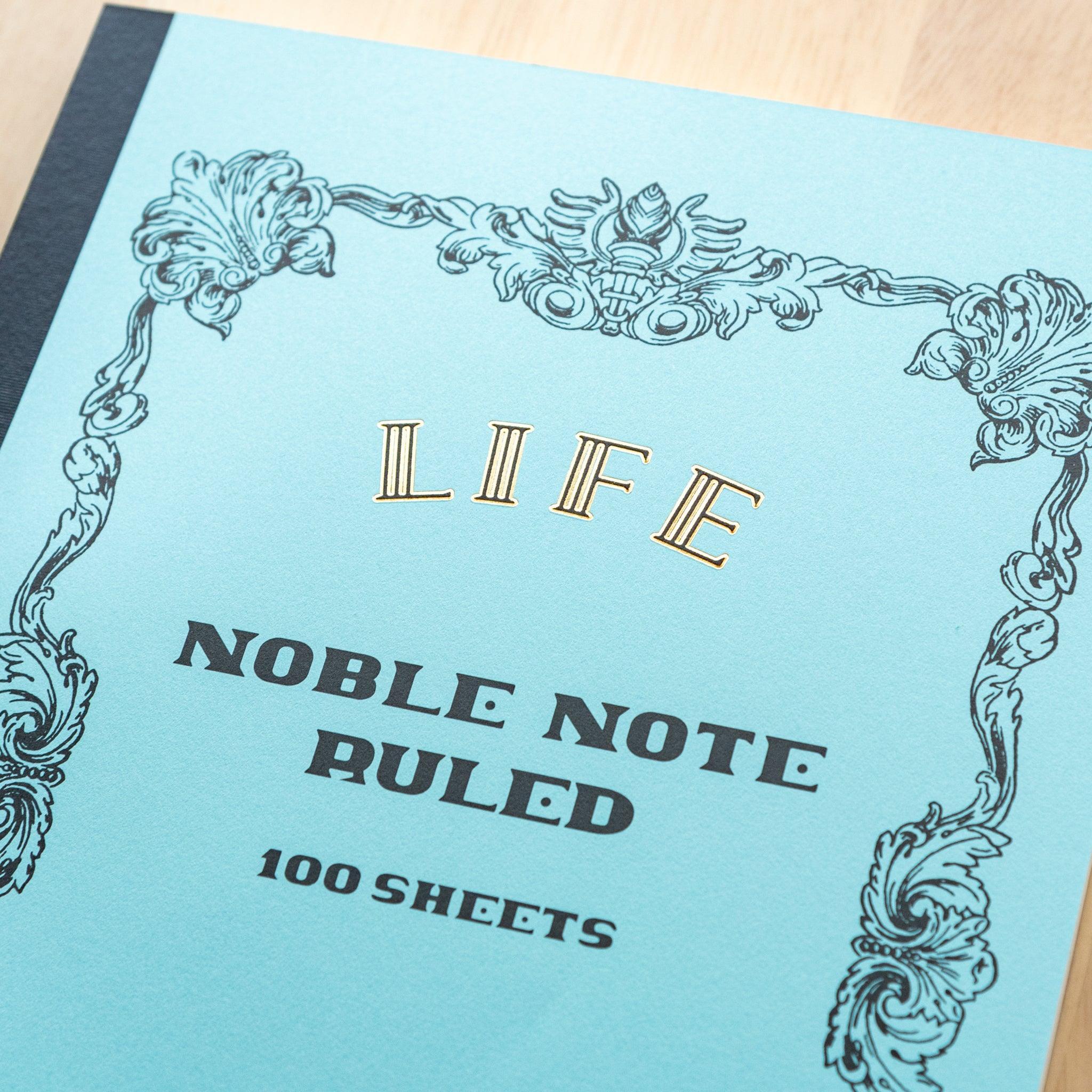 An A4-size LIFE Noble Note notebook with a pastel blue cover, gold lettering, and decorative black borders, labeled "Ruled, 100 Sheets."