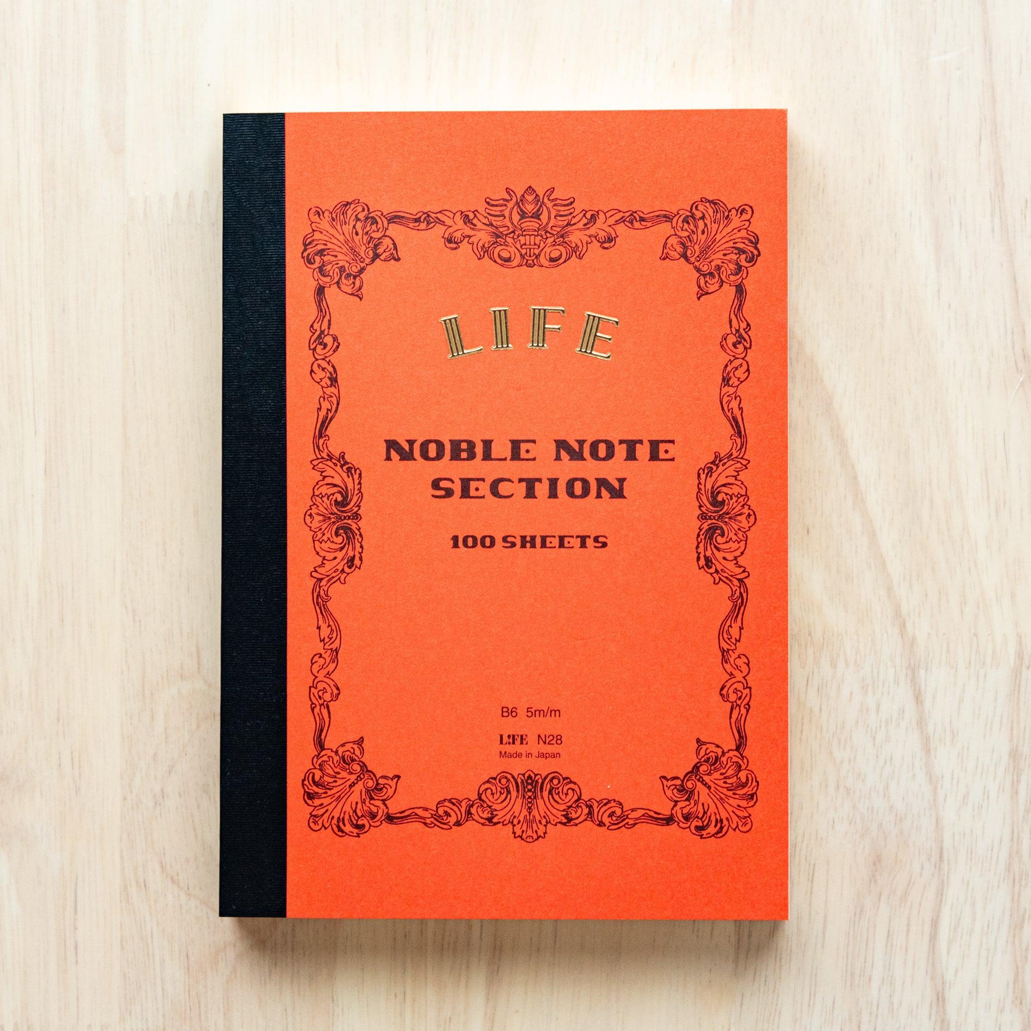 A single "LIFE Noble Note Ruled" notebook with a orange cover, black spine, ornate border design, and 100 sheets, displayed on a wooden surface.