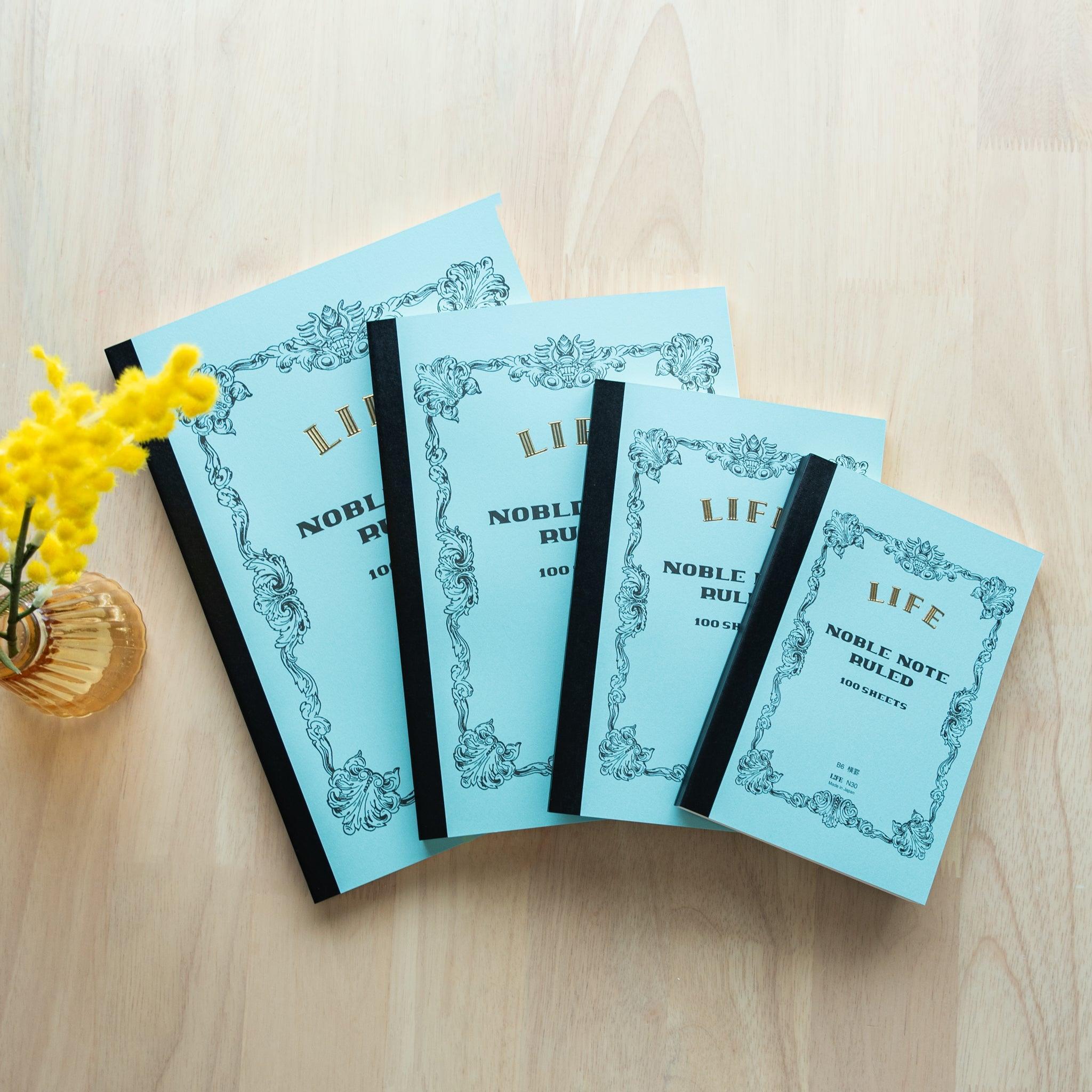 A set of blue "LIFE Noble Note Ruled" notebooks in four sizes with black spines and ornate designs, displayed on a wooden surface with yellow flowers.