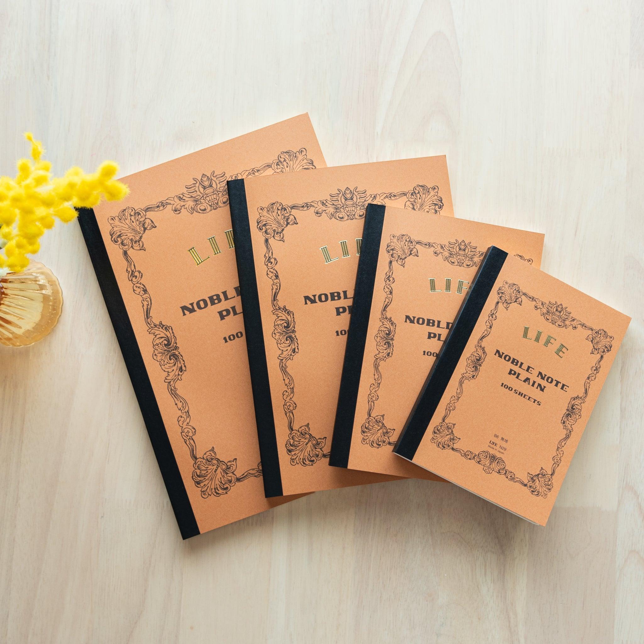 A set of brown "LIFE Noble Note Plain" notebooks in four sizes with black spines and ornate designs, displayed on a wooden surface with yellow flowers.