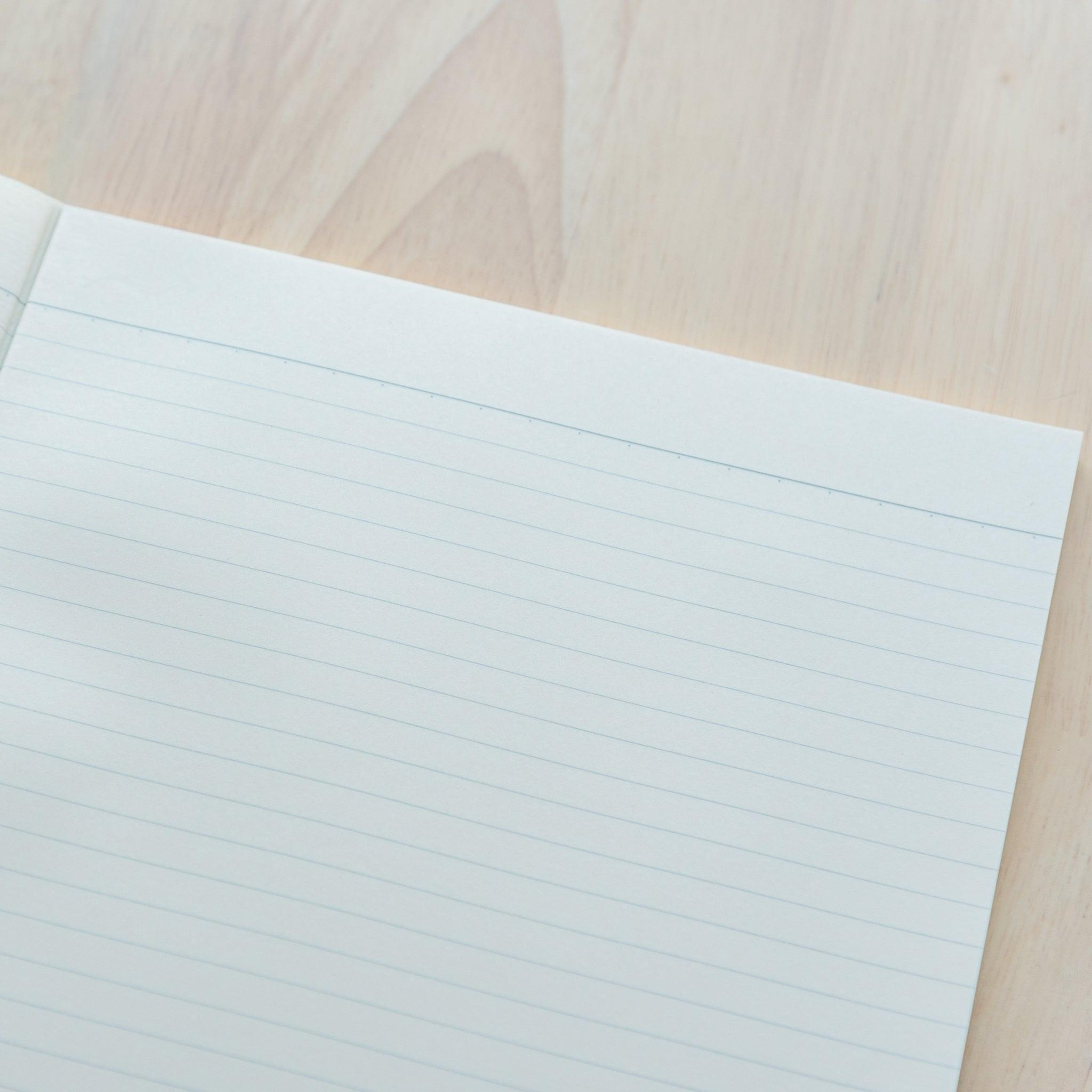 An open A4-size LIFE Noble Note notebook with cream-colored, lined pages and a smooth texture, placed on a wooden surface.