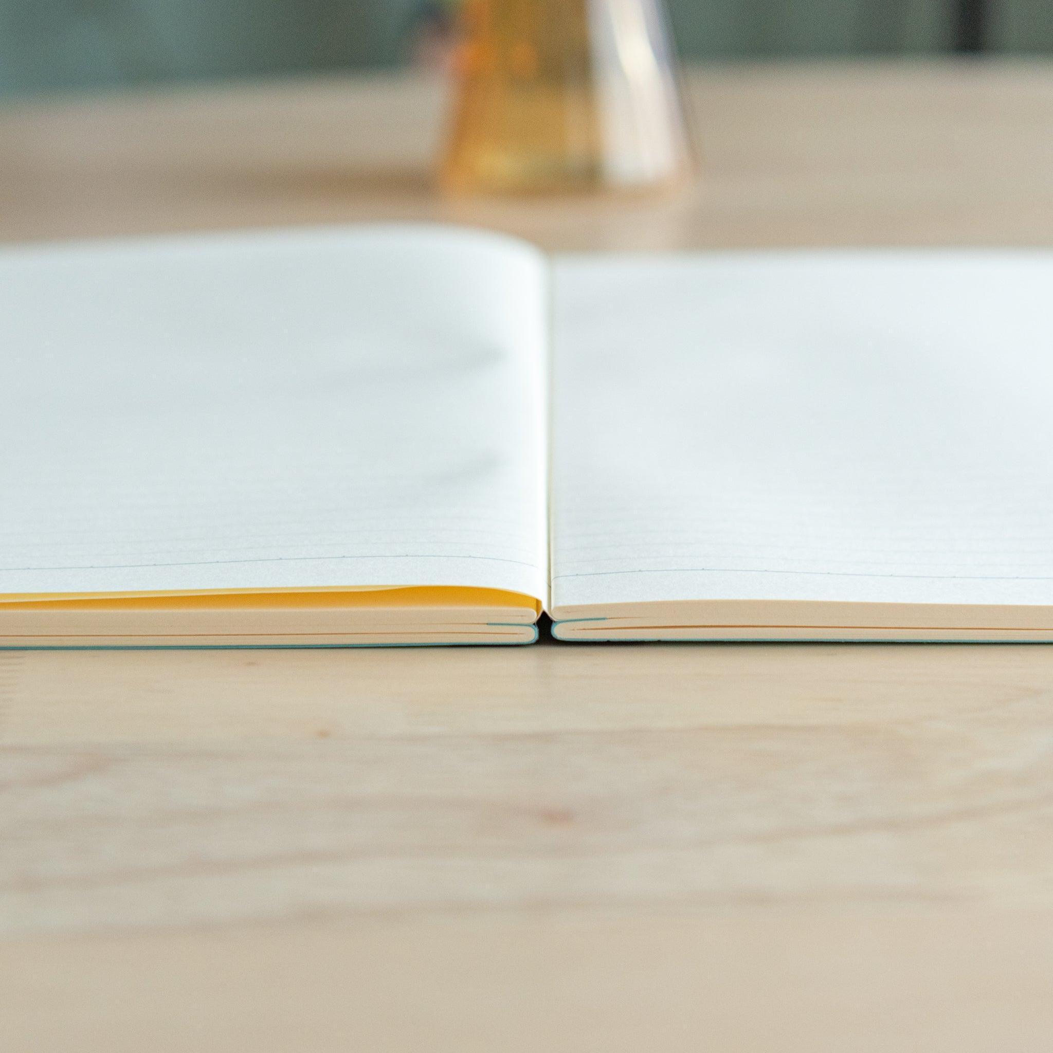 An open A4-size LIFE Noble Note notebook with cream-colored, lined pages and a smooth texture, placed on a wooden surface.