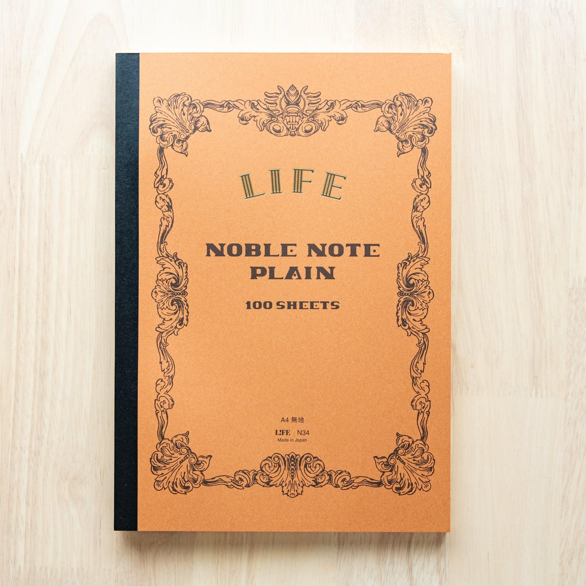 A single "LIFE Noble Note Plain" notebook with a brown cover, black spine, ornate border design, and 100 sheets, displayed on a wooden surface.