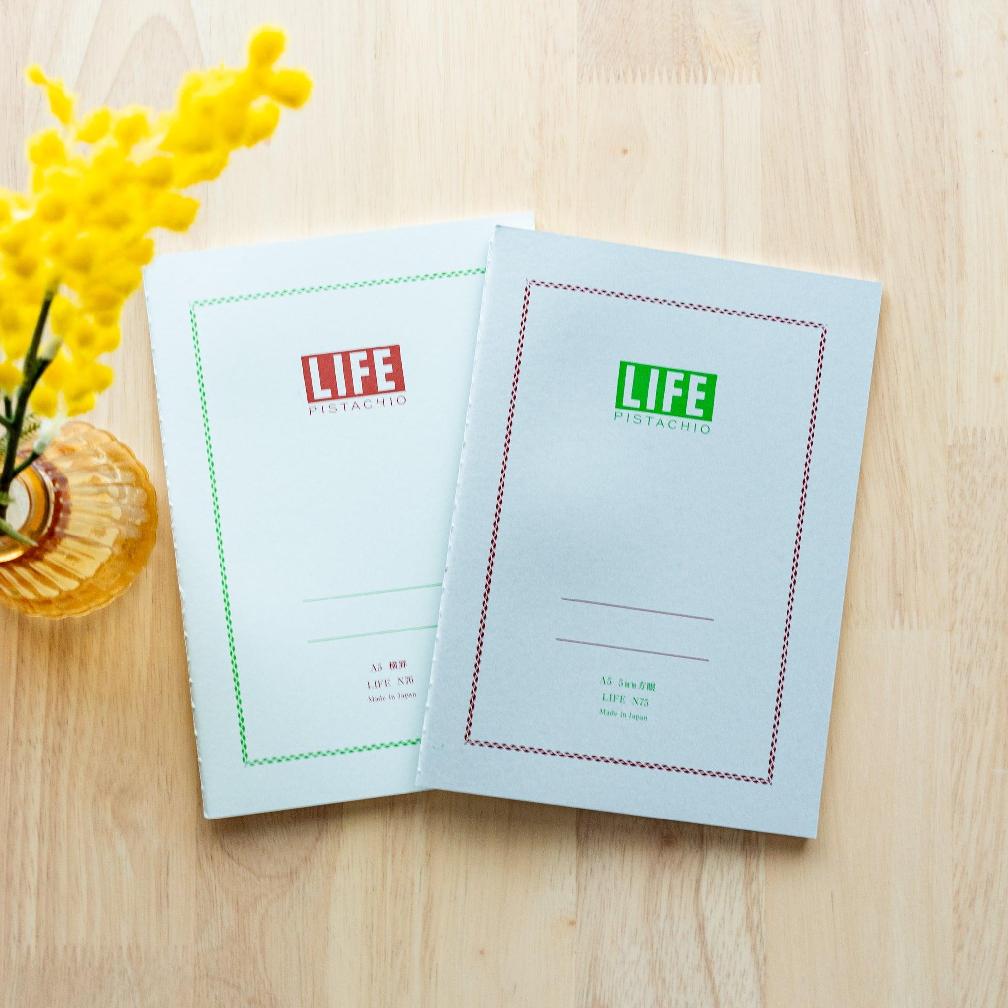 Two "LIFE Pistachio" notebooks in A5 size, one with a light gray cover and red logo, the other with green logo, displayed on a wooden surface with yellow flowers.