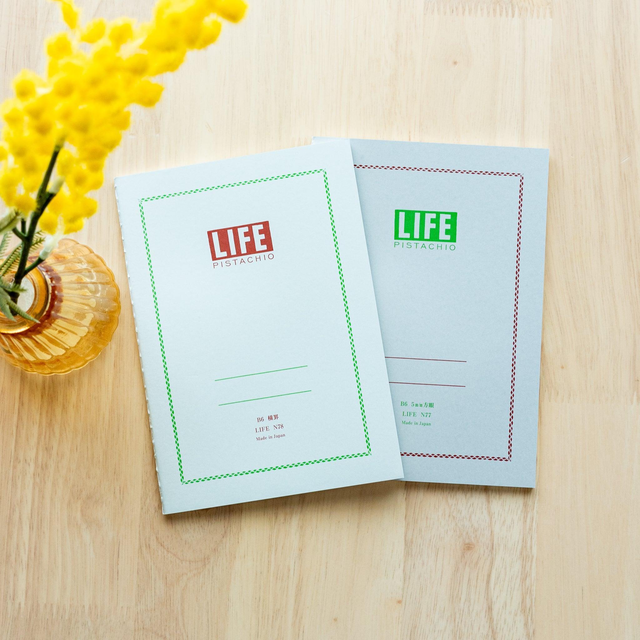 Two "LIFE Pistachio" notebooks in B6 size, one with a light gray cover and red logo, the other with green logo, displayed on a wooden surface with yellow flowers.