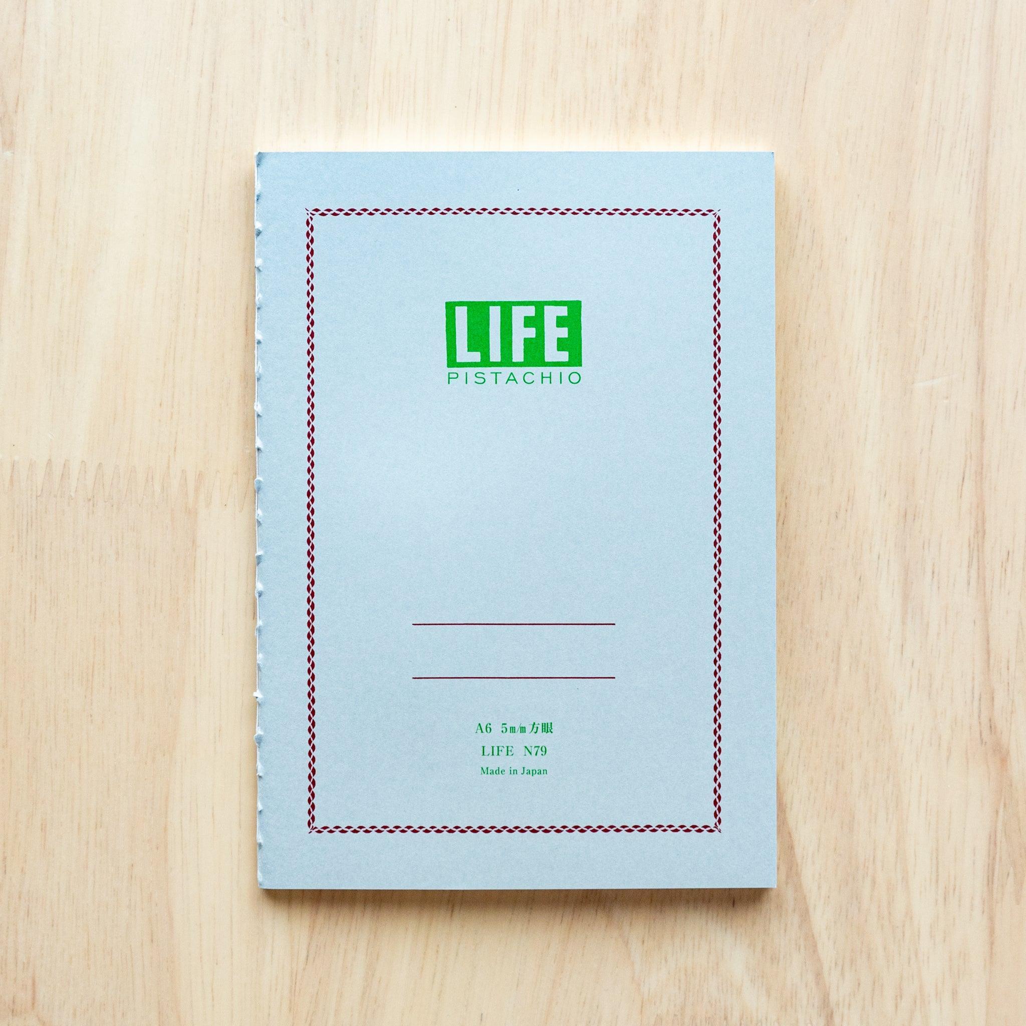 A single "LIFE Pistachio" notebook in A6 size with a light gray cover, green logo, and minimal border design, displayed on a wooden surface.