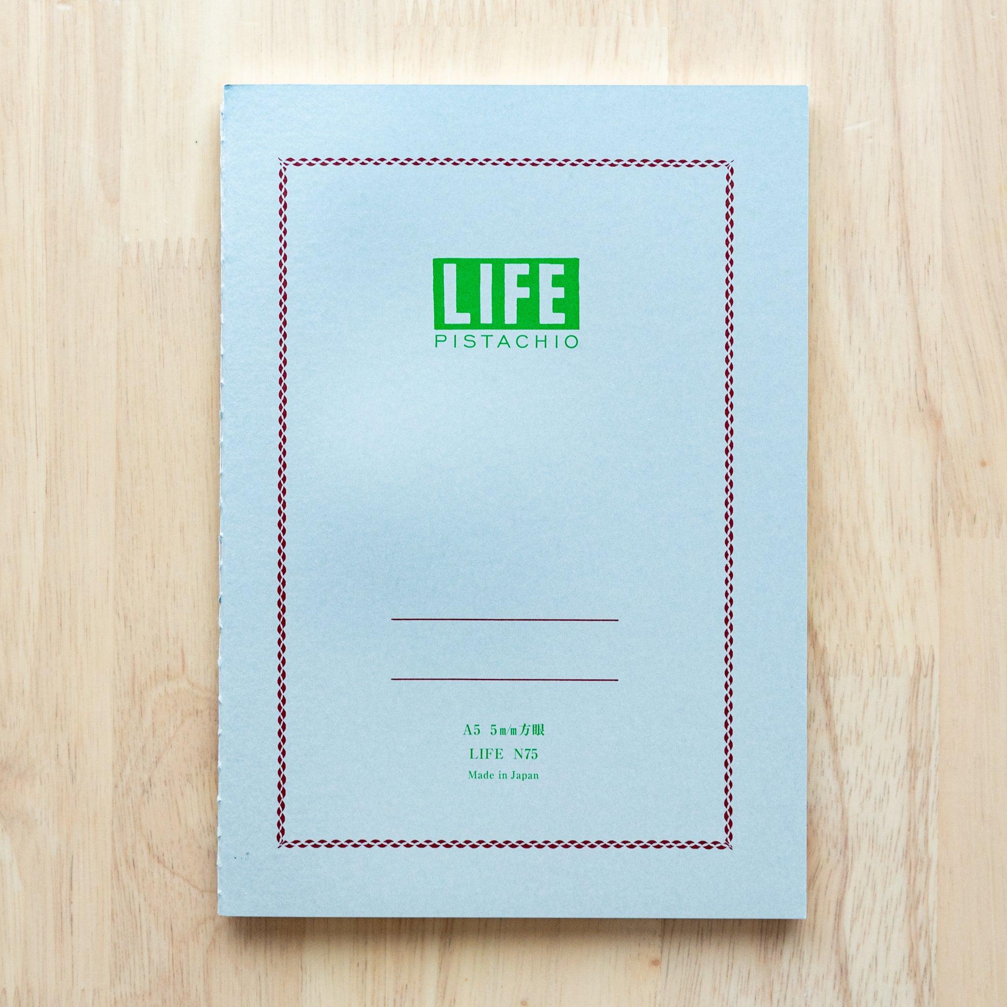 A single "LIFE Pistachio" notebook in A5 size with a light gray cover, green logo, and minimal border design, displayed on a wooden surface.