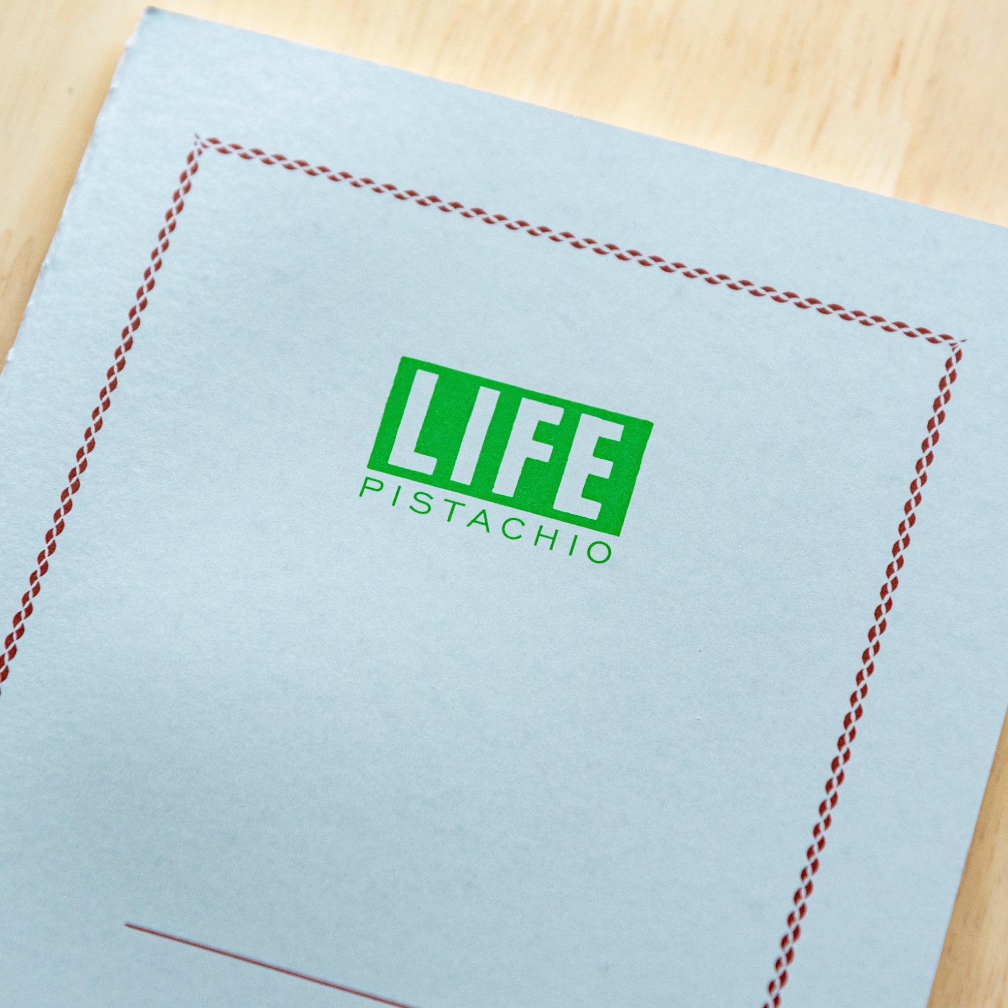A single "LIFE Pistachio" notebook in A5 size with a light gray cover, green logo, and minimal border design, displayed on a wooden surface.